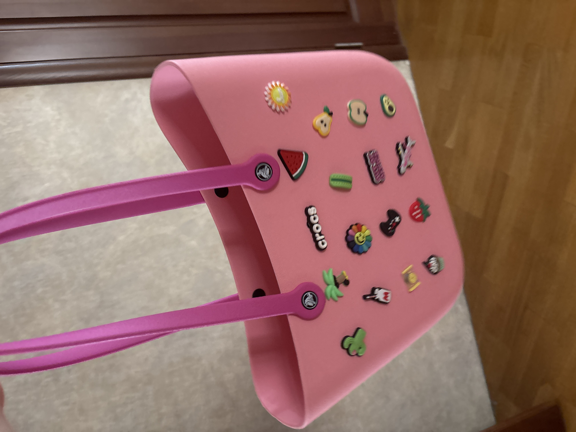 A close-up, slightly angled shot captures a pink Crocs tote bag adorned with various colorful charms. The bag features two pink handles and is decorated with charms such as a watermelon slice, a sun, a strawberry, an apple, an avocado, a pear, a cactus, a palm tree, a rainbow flower, and the word "Crocs". The background is blurred, showing a beige textured wall and a portion of a brown doorframe. The image appears to have been taken indoors. There is no identifiable information in the image to suggest a location in Georgiyevsk, Russia.
