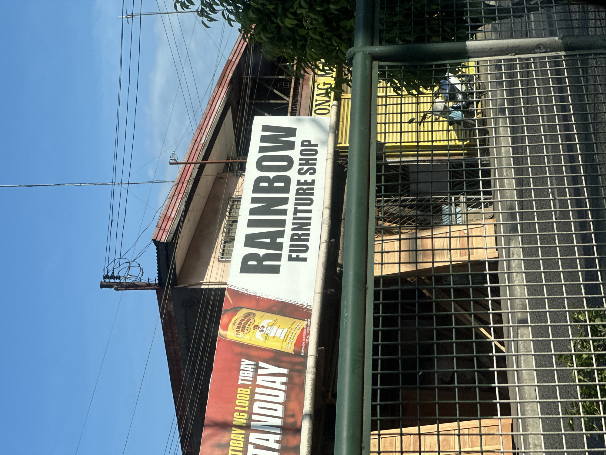 The image depicts a street scene in Davao City, Philippines, viewed from behind a dark green metal grid fence. In the immediate foreground, the fence casts shadows onto a concrete curb and dark asphalt road. Small green vegetation is visible below the fence line.
In the midground, a commercial area is visible, featuring a long billboard-style sign structure. The left section of the sign has a red background with white text "TIBAY NG LOOB. TIBAY TANDUAY" and a large yellow bottle of "TANDUAY RHUM DARK," with smaller print including "FOR 18+ ONLY. DRINK RESPONSIBLY. NSC NO. TDTZP120224T." The right section of the sign, on a white background, displays "RAINBOW FURNITURE SHOP" in large black lettering.
Behind the sign, parts of buildings are visible. To the left, an open storefront reveals stacked wooden planks or materials. To the right, a closed shop entrance features a bright yellow corrugated metal roll-down shutter, in front of which a light-colored scooter or motorcycle is parked. A partial sign with the text "ONAG M" is visible further to the right, behind the Rainbow Furniture Shop sign. The buildings above have red corrugated metal roofs and light-colored facades.
The background consists of a clear blue sky with minimal scattered white clouds. Numerous black electrical and communication wires are strung horizontally across the upper portion of the frame, connected to several visible utility poles. A tall, thin antenna-like pole extends upwards on the left, and a similar pole with a white tip is on the right. A portion of a green leafy tree or large bush is visible on the far right, partially obscuring a building.