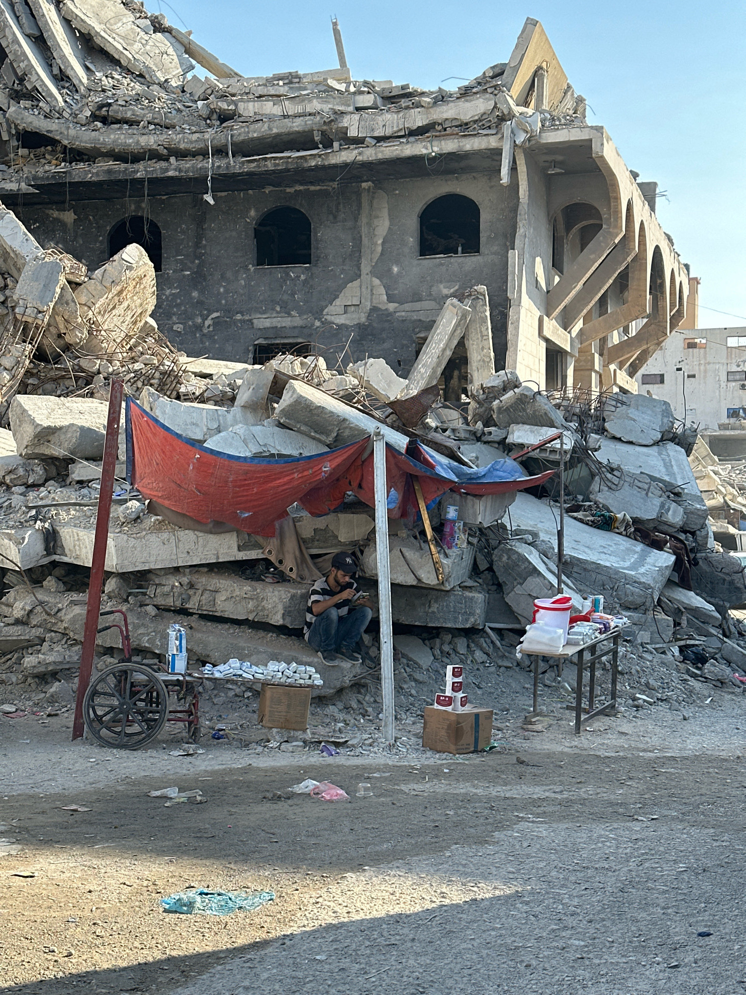 In Gaza City, Palestinian Territories, a man sits amidst the rubble of a destroyed building, using a blue tarp and a red sheet as a makeshift shelter. He is looking at his phone. In front of him, a cart with a single wheel is parked, and a small table displays various items, including what appear to be boxes of packaged goods and a red bucket. Piles of concrete debris dominate the background, with the skeletal remains of the building still visible. The ground is uneven and littered with debris.