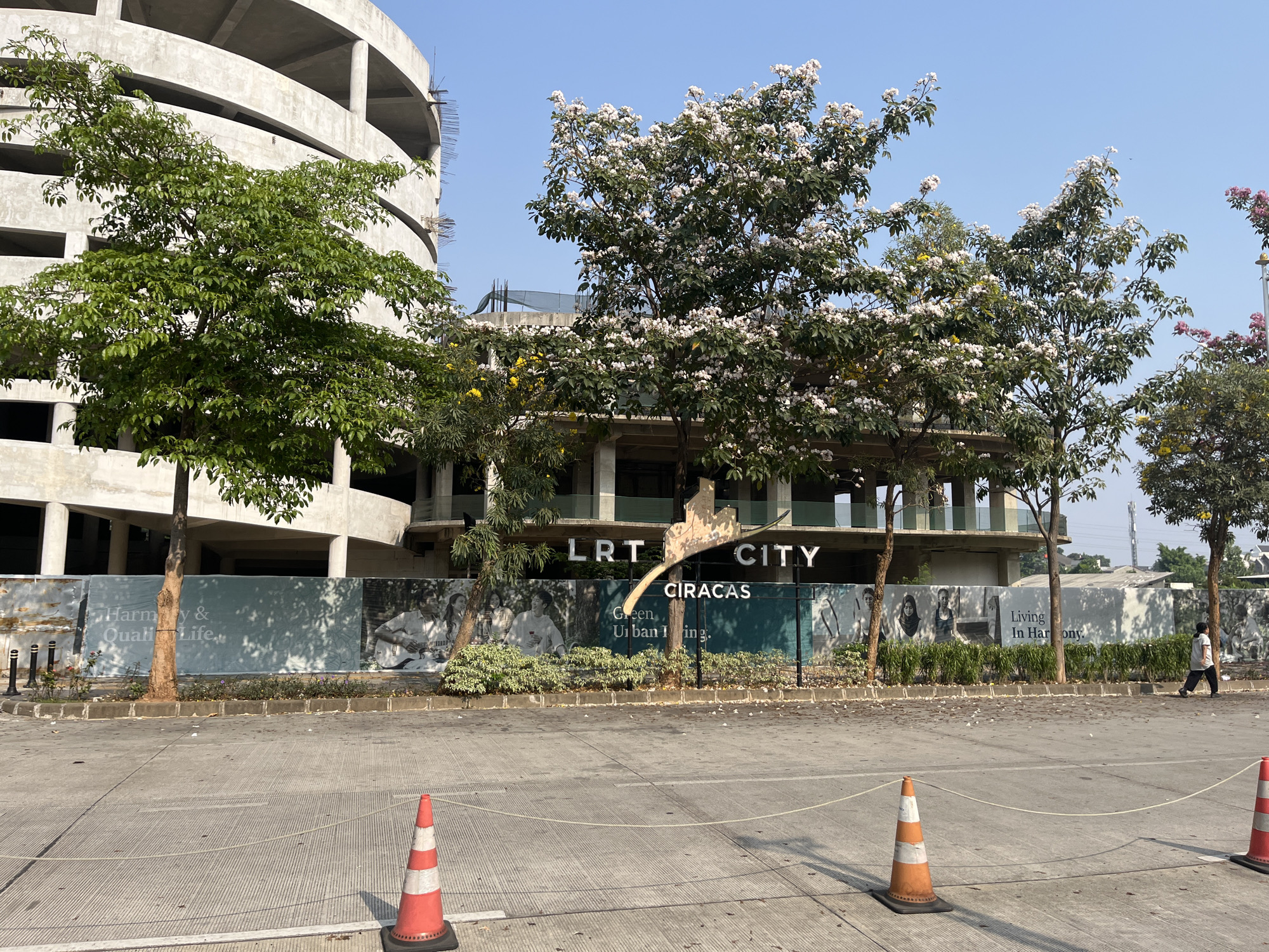 A construction site is visible in East Jakarta, Indonesia, with a partially built concrete building on the left and a modern building with glass balconies in the center. Trees with white and yellow flowers are in the foreground, casting shadows on a large banner that reads "LRT CITY CIRACAS" and "Green Urban Living." There is a person walking on the right side of the image. The ground is paved with concrete and marked with lines, with orange traffic cones and a white rope barrier in the foreground. The sky is clear and blue.