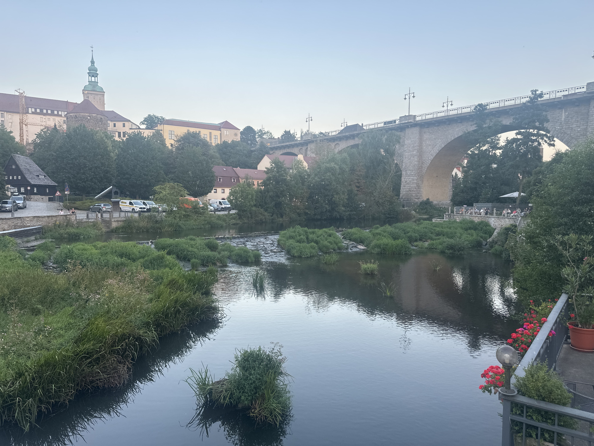 This image depicts a view of the Spree River in Bautzen, Germany, looking towards the historic Ortenburg Castle complex and the iconic Stone Bridge. The Ortenburg Castle, with its prominent green-spired tower and stone fortifications, sits atop a hill on the left side of the frame, surrounded by trees and other buildings. The Stone Bridge, a massive stone arch structure, spans the river in the upper right quadrant of the image, carrying a road and pedestrian walkway. Several cars are parked along the riverside road below the castle. The river itself is calm and reflects the sky and surrounding greenery. Patches of lush green vegetation, including reeds and small islands, dot the water. In the foreground on the right, a section of a balcony or terrace is visible with potted plants, red flowers, and a vintage-style lamp. Several people are seated at tables on the terrace. The sky is overcast and a pale blue, suggesting either early morning or late evening. The overall scene conveys a peaceful, historic urban landscape.