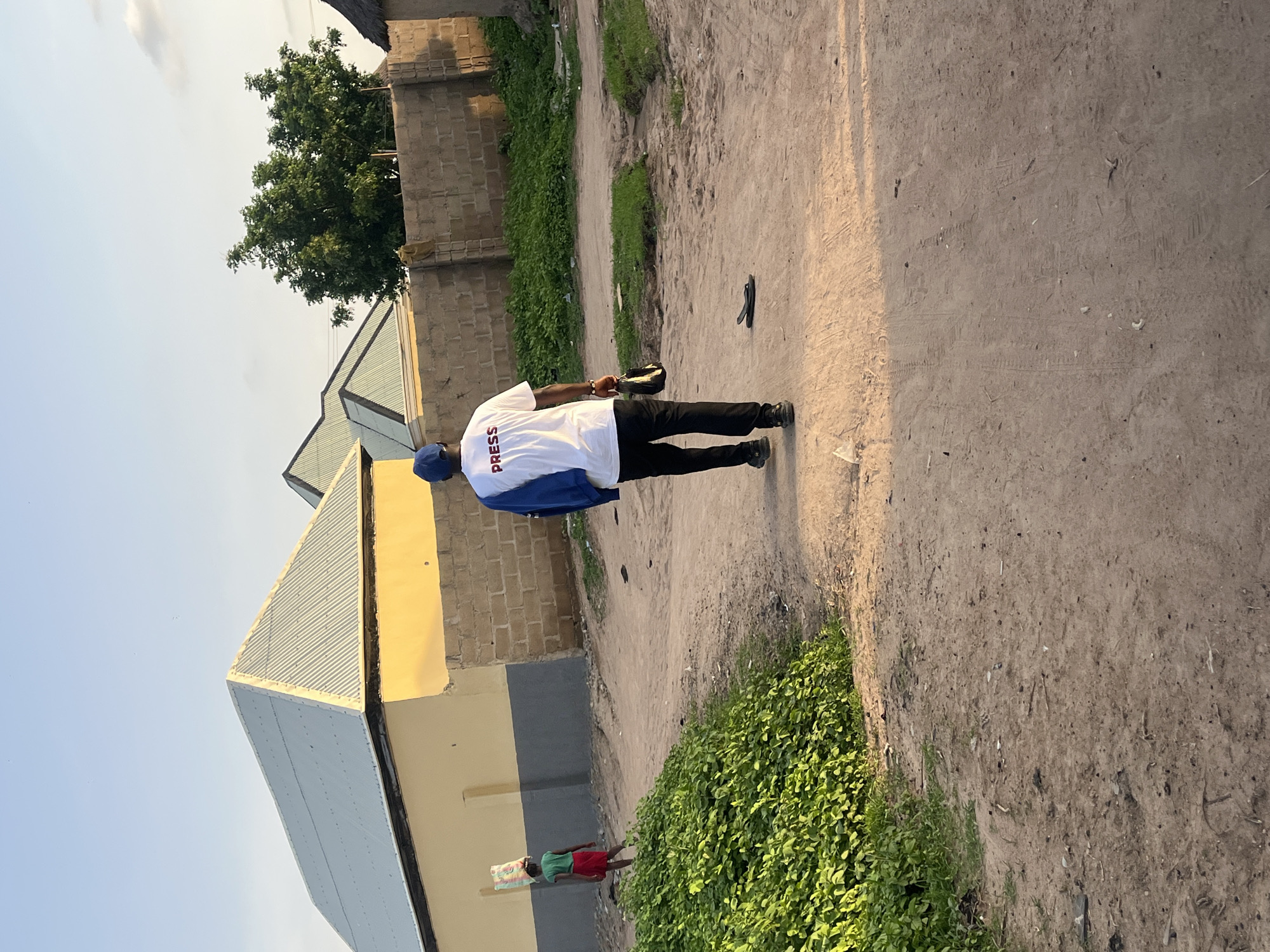 In Jalingo, Nigeria, a man wearing a white t-shirt with the word "PRESS" in red, black pants, a blue cap, and a blue jacket draped over his shoulder walks away from the camera on a dirt road. He is carrying a black bag in his right hand. To his left, a young girl in a green t-shirt and red skirt walks away from the camera, carrying a large bag of goods on her head. The environment is a residential area with buildings constructed from yellow and grey blocks, and a brick wall topped with a thatched roof. Lush green vegetation borders the road on the left. The sky is clear and a soft golden light indicates it is either early morning or late afternoon.