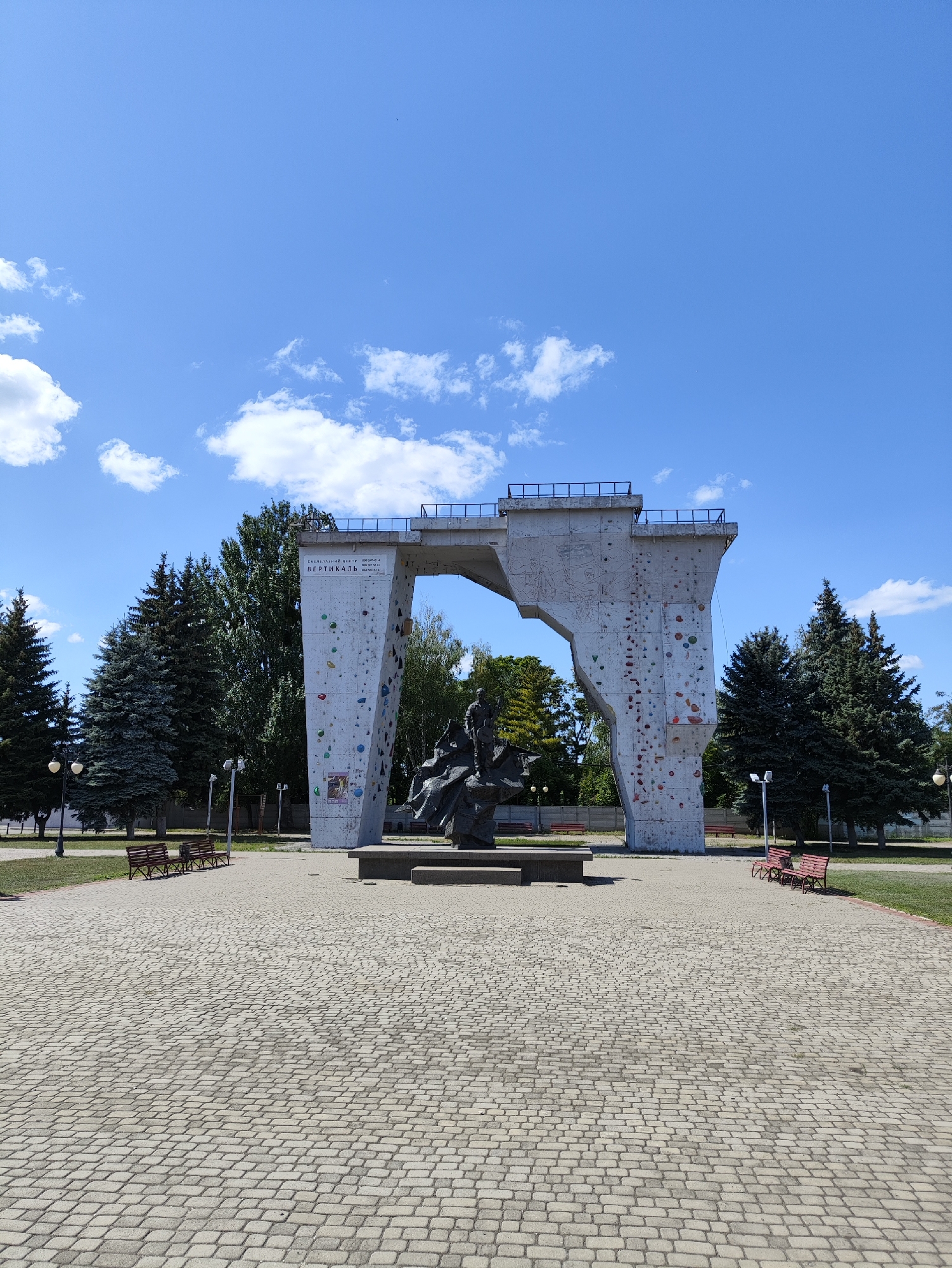 The image depicts a large concrete structure resembling an archway, with one side designed as a climbing wall. The climbing wall features numerous colorful holds. In the foreground, a paved area with interlocking stone bricks leads towards the structure. A dark bronze statue of a seated figure is positioned in front of the climbing wall. Several empty red benches are visible on a grassy area to the left and right of the paved path. Tall evergreen trees and deciduous trees form a backdrop behind the structure. The sky is bright blue with scattered white clouds. The scene is set in a public park or recreational area. This location is known as the "Vertical" climbing wall in Kharkiv, Ukraine.