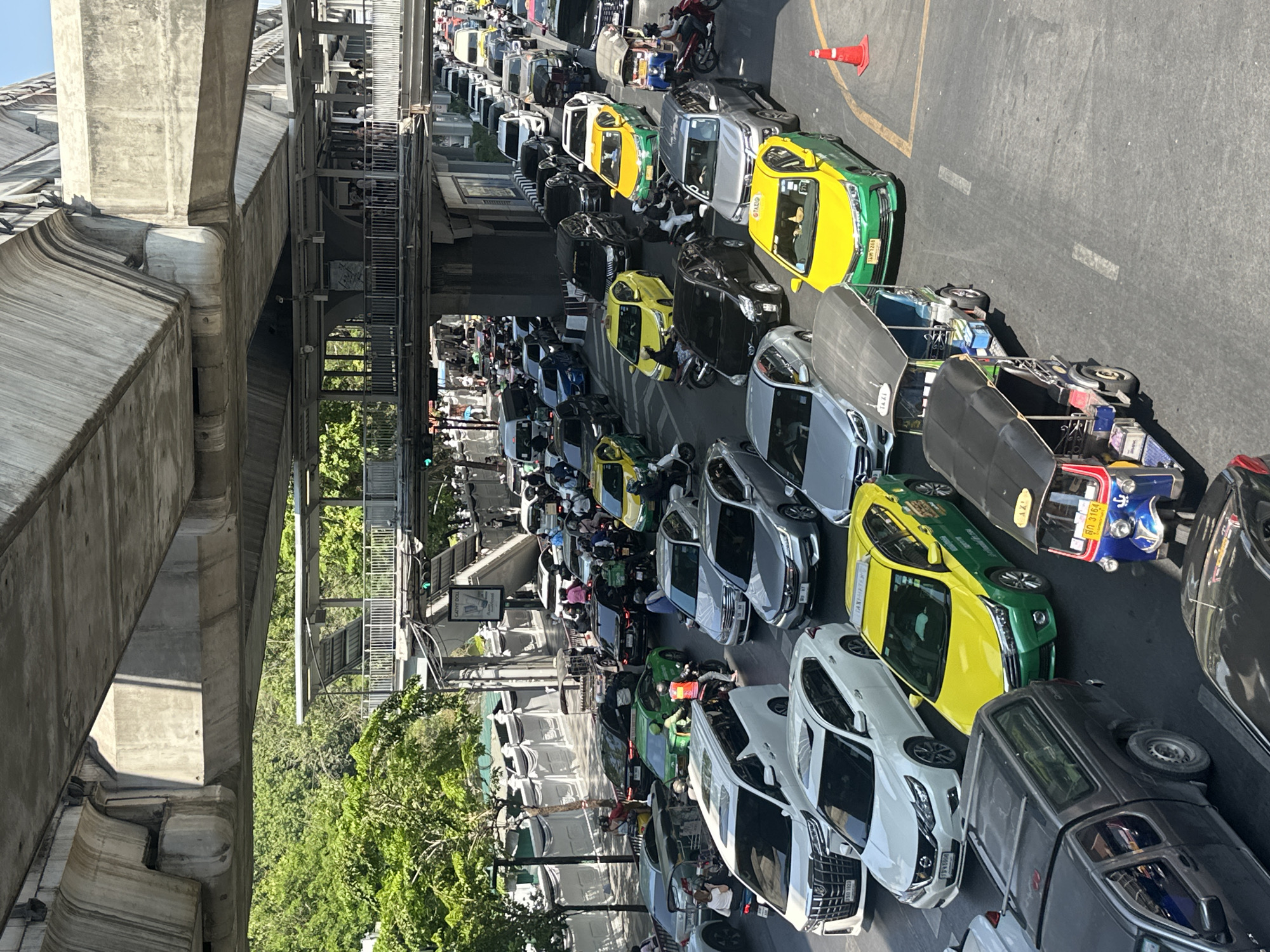 The image depicts a traffic jam in Bangkok, Thailand. The location is likely in a densely populated area, indicated by the elevated walkways and overhead structures visible. The traffic consists of a mix of vehicles, including cars, motorcycles, and a tuk-tuk. There are many yellow and green taxis intermingled in the traffic, consistent with the color scheme of Bangkok's taxi fleet. Pedestrians can be seen walking on the sidewalks and elevated walkways, suggesting a busy urban environment. The light conditions indicate it is daytime, with a clear sky. An orange traffic cone is visible on the right side of the image.
