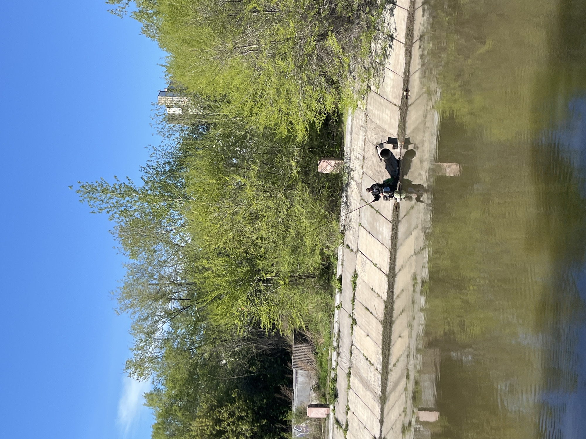 The image depicts a section of a concrete canal embankment in Krasnoyarsk, Russia, with water on the right side and trees and vegetation on the left. The sky is blue. Two people are fishing on the embankment. There is a building visible behind the trees. A small, dilapidated structure sits at the base of the trees.