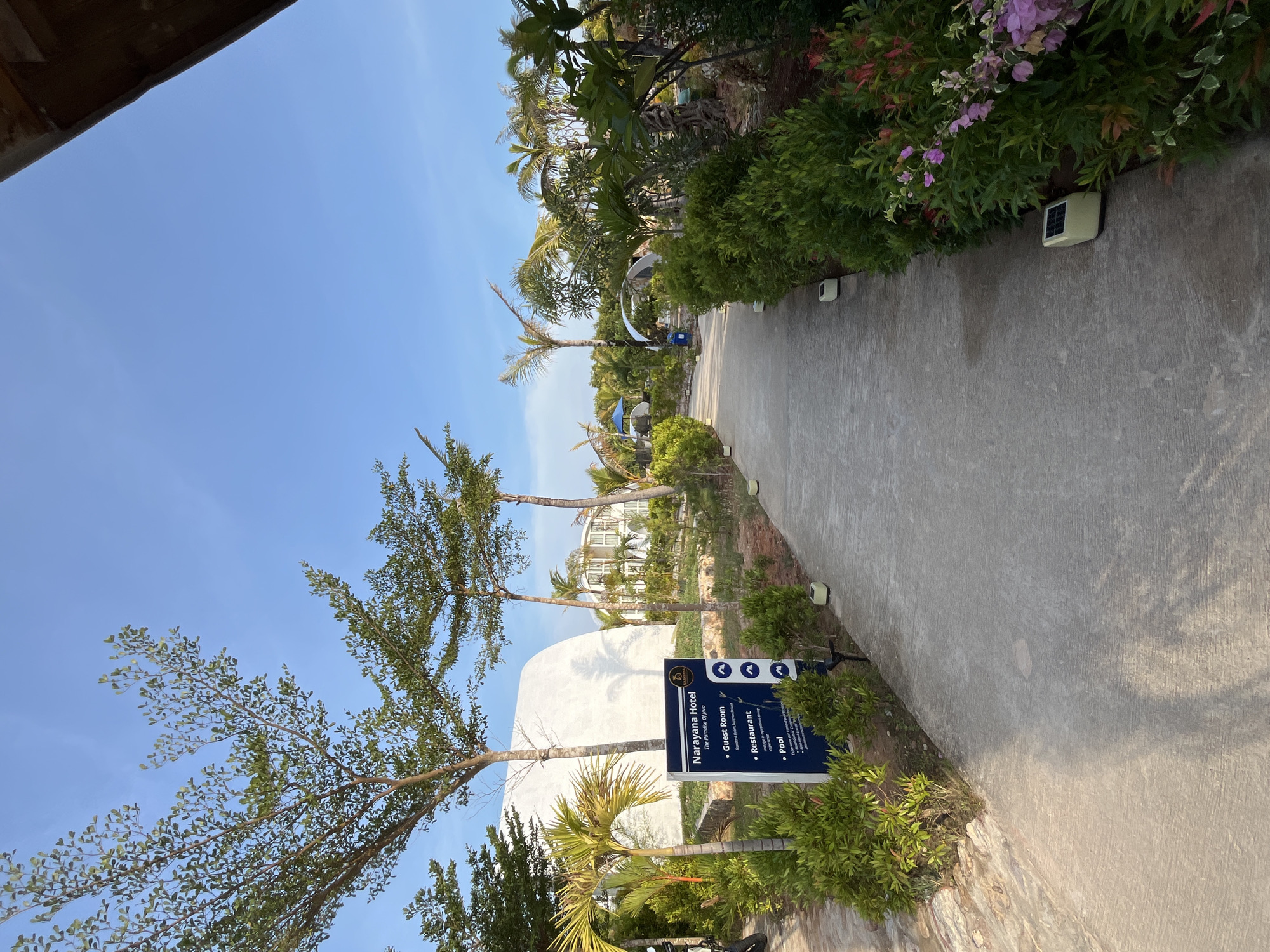 This is a landscape photograph taken in Kemloko, Indonesia. The scene depicts a pathway leading into a resort or hotel complex. On the left side of the pathway, a sign indicates "Narayana Hotel" and lists various amenities such as "Guest Room," "Restaurant," and "Pool." The pathway is paved with concrete and lined with lush green foliage and small trees. In the background, there are white dome-shaped structures, palm trees, and other tropical vegetation. The sky is clear and blue, suggesting a sunny day. The overall impression is one of a serene and tropical vacation destination.