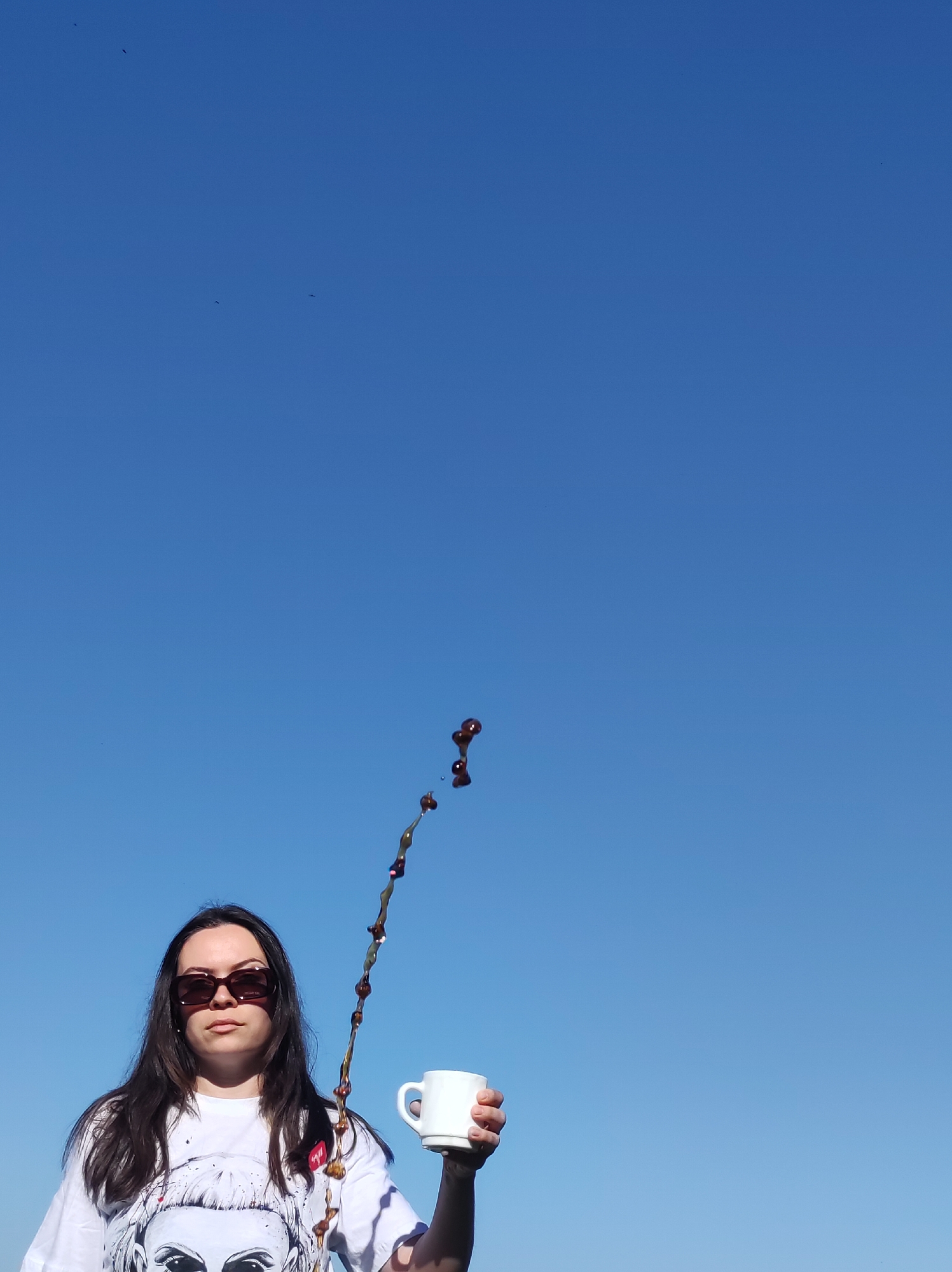 A woman in sunglasses stands against a cloudless blue sky, holding a white mug from which a dark liquid (likely coffee) is splashing upwards. She is wearing a white t-shirt with a design printed on it. The location is not discernible from the image. There is no data to suggest the location is Фоки, Russia.