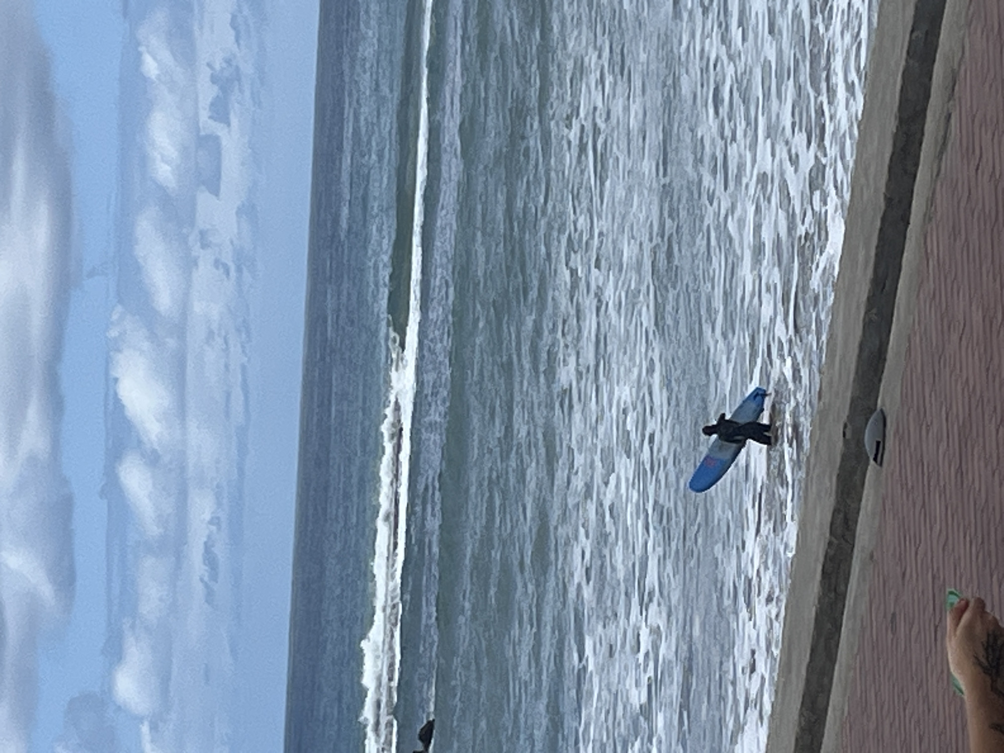 The image displays an ocean scene on a bright, clear day. A person, dressed in dark attire, is visible lying on a blue surfboard in the foamy, light-grey water, seemingly paddling or waiting for a wave. The ocean dominates the mid-ground, characterized by rolling waves, some breaking into white surf closer to the foreground, while others appear as calmer swells further out.
To the left, the sky is a soft blue with scattered, fluffy white clouds. To the right, a concrete seawall or promenade runs along the water's edge. This structure consists of a lighter grey, smooth concrete surface adjacent to the sea, backed by a darker, reddish-brown tiled or textured wall. A small, grey, semi-circular fixture is attached to the concrete wall.
In the bottom right corner of the frame, a human foot with green-painted toenails and a portion of a tattooed arm or hand are visible, holding a small green object. This suggests another individual is observing the scene from the promenade. The overall impression is one of a coastal setting under pleasant weather conditions, likely during daytime. No text is discernible in the image.