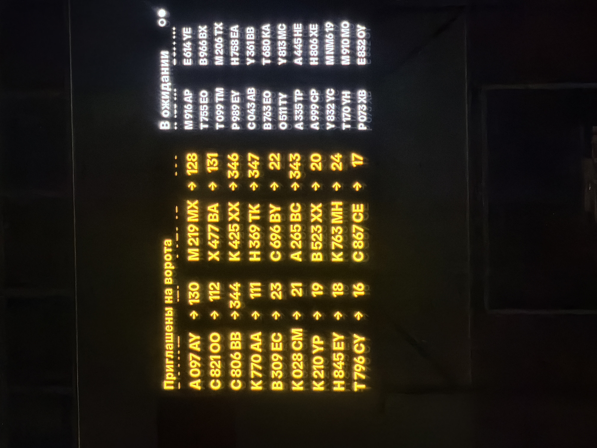 The image shows a digital display board with yellow and white text on a black background. The text is in Russian and English. The top of the display board has the Russian text "Приглашены на ворота", which translates to "Boarding at the gates". Below this, there are lists of codes and numbers, some with arrows, suggesting flight or gate information.
To the right of the yellow text, there is a section with white text that reads "В ожидании", which translates to "Waiting". This section also lists codes and numbers.
Based on the visual cues of a digital display board commonly found in airports, and the Russian text, this image likely depicts a departures or arrivals board at an airport. Without further information or context, it is not possible to definitively determine the specific location within Sluchainyi, Russia. However, such displays are characteristic of airport terminals.
The subjects are the text on the display board. The objects are the digital display board itself. The environment is likely an indoor public space, possibly an airport terminal. There are no people directly visible in the image, only the information presented on the screen. The lighting appears to be dim, with the display board being the primary light source in its immediate vicinity.