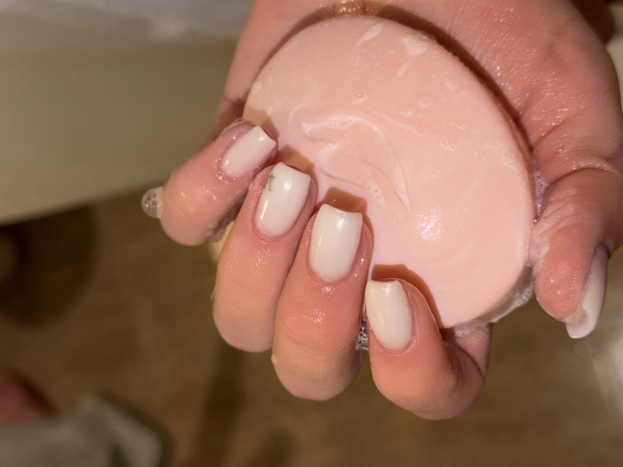 A close-up, high-angle shot depicts a wet human hand, likely female, actively holding and lathering a bar of soap. The hand's skin is moist, with visible water droplets and white soap suds. The fingernails are manicured with an opaque, light-colored polish. The ring finger's nail features a small, dark, linear mark or design. The object held is a pink, oval, or circular bar of soap, also covered in white lather and moisture. Subtle embossed patterns are discernible on the soap's surface beneath the suds. The background is an out-of-focus indoor environment, consisting of blurred light brown and off-white tones, consistent with a washroom or bathroom. This scene occurs in Sofia, Bulgaria, within an unidentifiable specific indoor location.