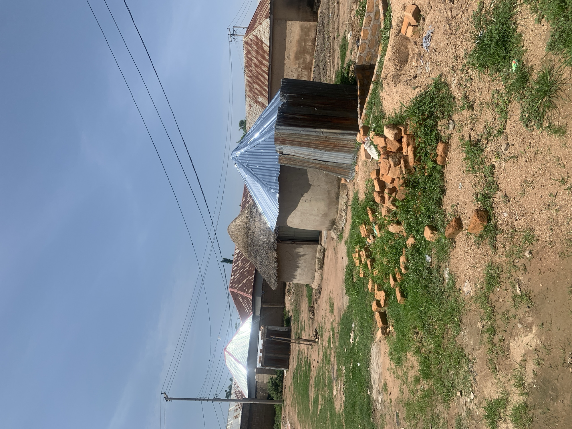 In the city of Jauro Yinu, Nigeria, a rural scene unfolds under a clear blue sky with scattered, wispy clouds. The foreground is dominated by a patch of uneven, dry earth covered with sparse green grass and a pile of rough, red-brown bricks. Further back, a dirt path meanders towards a cluster of round, mud-walled huts. Two of these huts have conical roofs, one thatched with dried grass and the other covered with corrugated metal, shimmering in the sunlight. Adjacent to these, a larger, rectangular building features a similarly corrugated metal roof, weathered and reddish-brown. Another building with a more elaborately designed corrugated metal roof, appearing lighter and brighter, is also visible. A low, brick wall demarcates a construction area in the mid-ground. A wooden structure, possibly a shed or storage area, stands between two of the huts. Electrical wires crisscross the sky, supported by a utility pole on the left side of the frame. The overall impression is one of a developing rural settlement, with a mix of traditional and modern building materials.