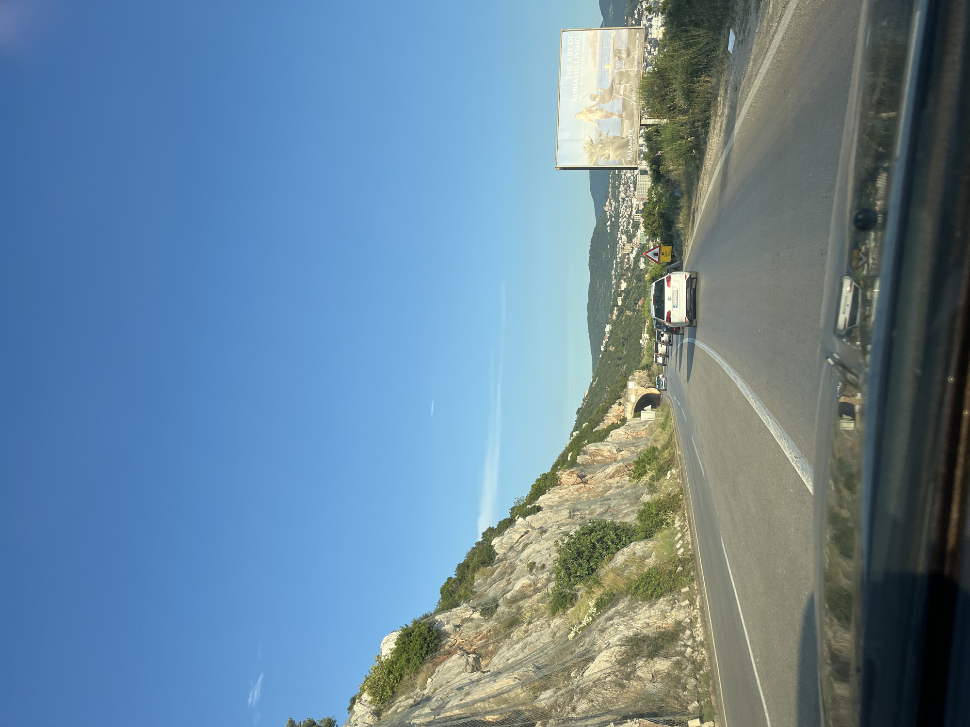 The image captures a scenic view of a coastal road in Dobra Voda, Montenegro, on a clear, sunny day. The perspective is from inside a vehicle, looking forward. The road curves to the right, with a steep, rocky mountainside on the left, reinforced with netting to prevent rockfalls. Vegetation, including trees and shrubs, grows on the mountainside and along the roadside. A tunnel entrance is visible in the distance on the right side of the road. Several cars are traveling on the road, with a white SUV prominently in the foreground. On the right side of the road, a large billboard advertises "THE ART OF ADRIATIC LIVING" with an image of people relaxing by the sea, and mentions "KALAMPER HOTEL & SPA". In the background, a town with buildings is nestled on the hillsides, overlooking the Adriatic Sea. The sky is a vibrant, cloudless blue.
