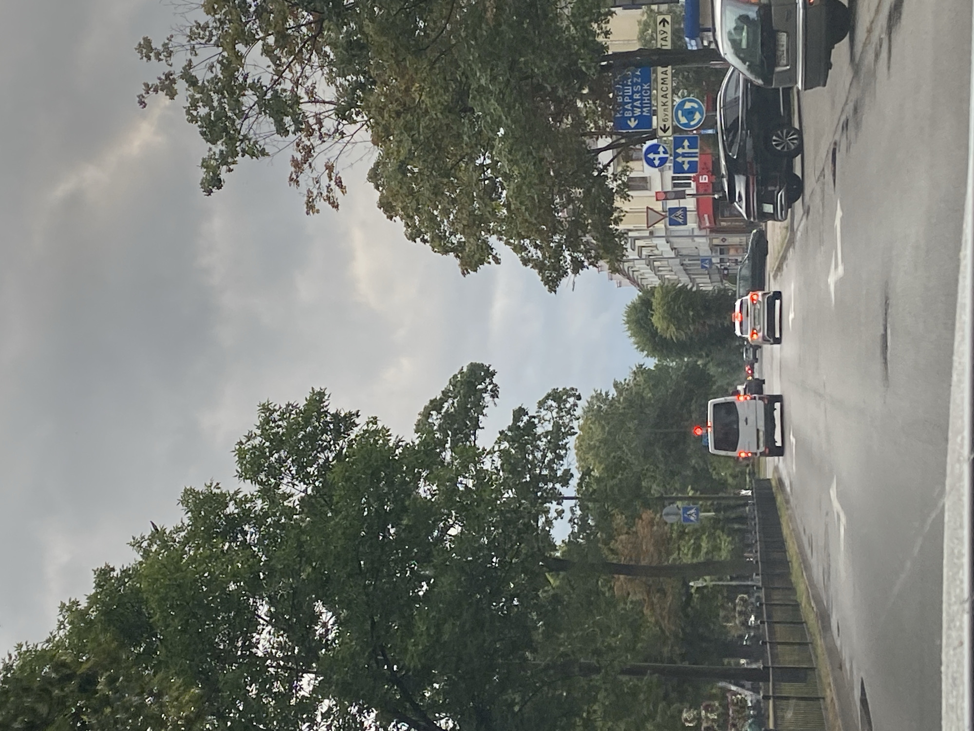 The image depicts a view from a vehicle driving on a wet, asphalt road in the city of Brest, Belarus. The sky is overcast with gray clouds. The road is lined with trees on both sides, and buildings are visible in the distance. Several cars are on the road, including a white van with its brake lights illuminated. Traffic signs are present, indicating directions to Minsk and Warsaw. The scene appears to be a typical urban street with moderate traffic under cloudy weather conditions.
