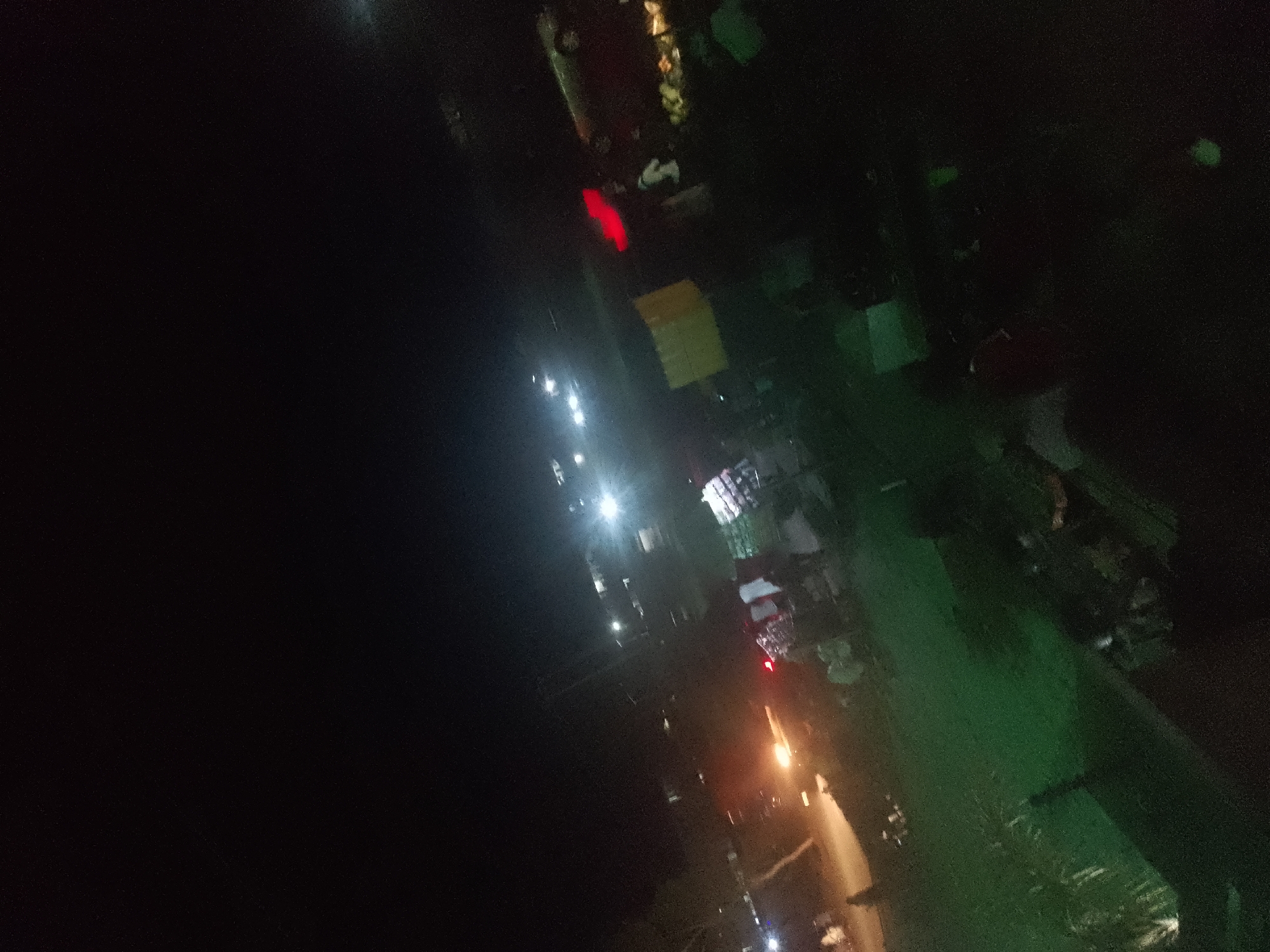 The image captures a dimly lit scene in Jalingo, Nigeria, at night, likely a street or outdoor market area. The composition is dominated by darkness, with scattered points of artificial light illuminating various elements. In the upper central area, a prominent red light is visible, possibly from a vehicle or signage. Below this, a bright yellow rectangular object is discernible. Further down, several white streetlights cast a glow on what appears to be a paved surface or roadway, with a blurred indication of buildings or structures on the left. On the right side of the image, a swath of green suggests vegetation or a grassy area, interspersed with darker shapes that could be objects or people. Orange and white lights from lamps or vehicles are present towards the bottom right, illuminating a pathway or road. The overall impression is of a somewhat chaotic or busy nocturnal environment, typical of a developing urban area in Nigeria.