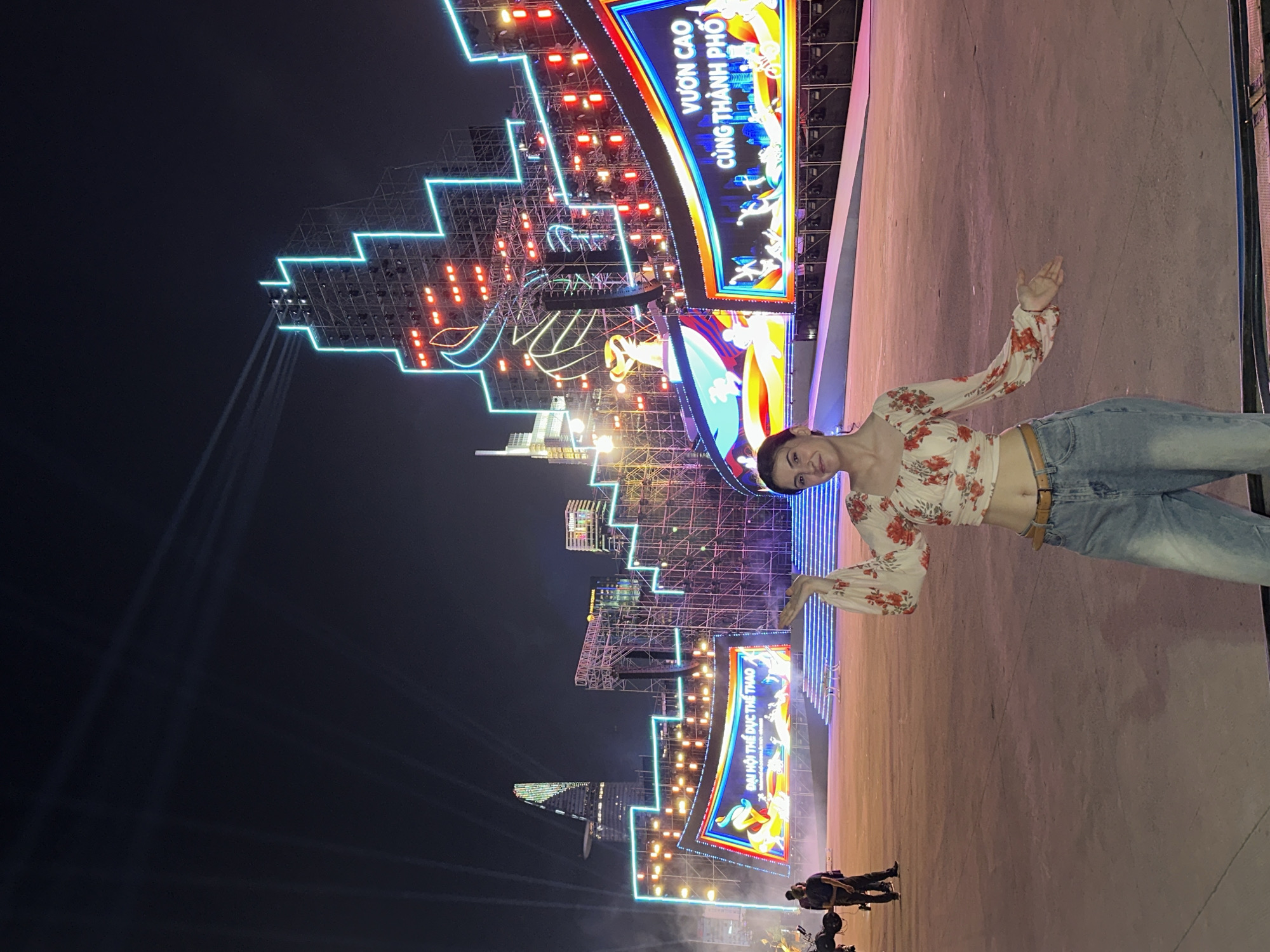A medium shot shows a slender fair-skinned woman with shoulder-length dark hair in front of an illuminated stage at night in the city of Thủ Đức, Vietnam. The woman wears a cropped, long-sleeved, light-colored blouse with a floral pattern and light-wash jeans. She extends both hands with open palms, gesturing toward the stage.
The stage displays tiered, illuminated scaffolding, with glowing neon outlines in blue and red, suggesting a cityscape silhouette. Large, colorful signs with Vietnamese script are mounted on the scaffolding. Spotlights shine upward into the night sky. In the foreground, smaller figures of individuals and equipment are discernible at the base of the stage. The ground surface appears to be concrete.