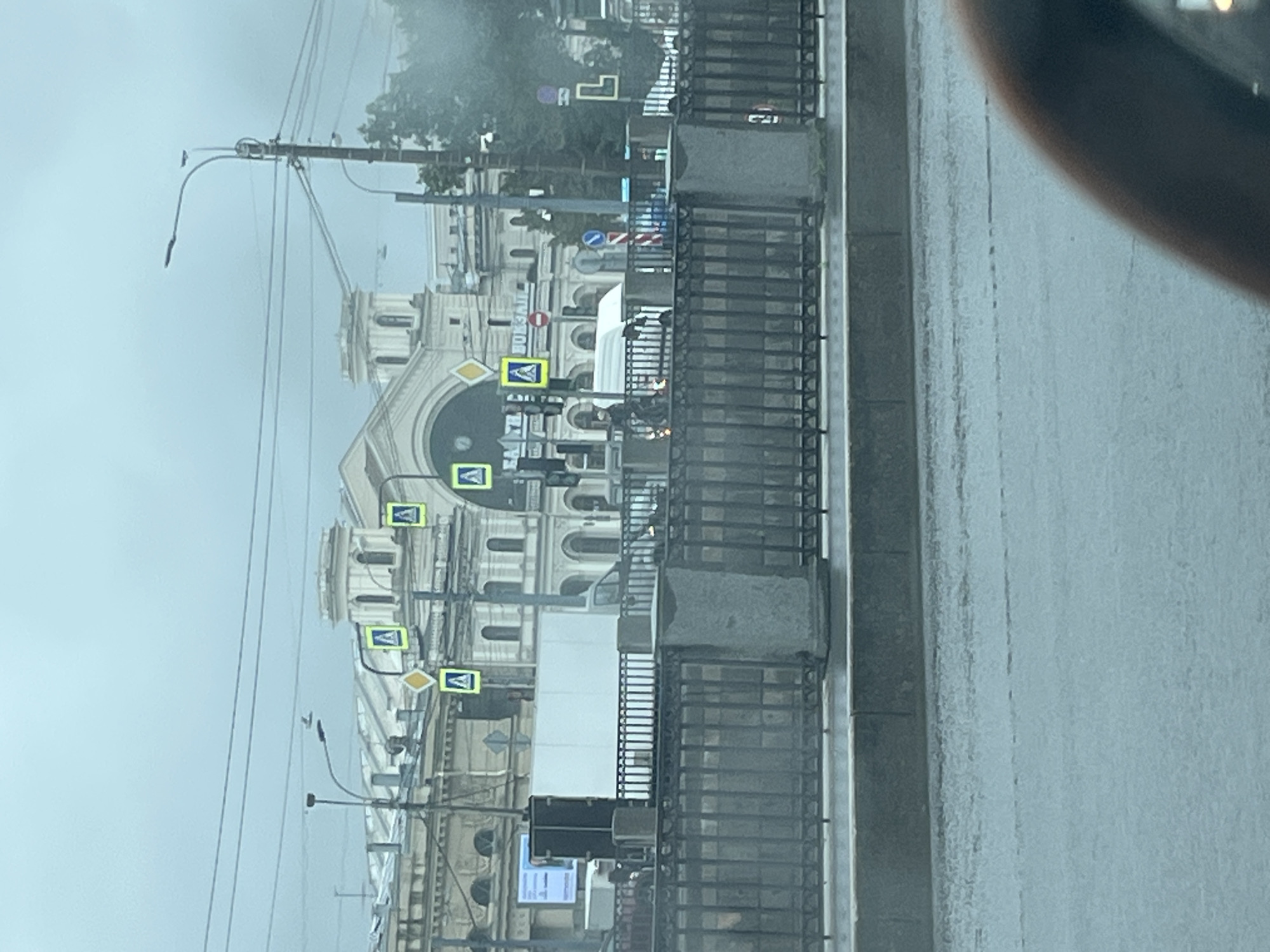 The image shows a view of the Moskovsky Railway Station in Saint Petersburg, Russia, on a cloudy day. The station's grand facade, with its classical architecture, is visible in the background. In the foreground, a black metal fence runs across the width of the image, separating the street from a sidewalk or pedestrian area. Beyond the fence, a white truck is parked. Several yellow pedestrian crossing signs are mounted on poles. The street in the immediate foreground is wet, suggesting it has recently rained. Part of a car's interior is visible in the bottom right corner of the frame.