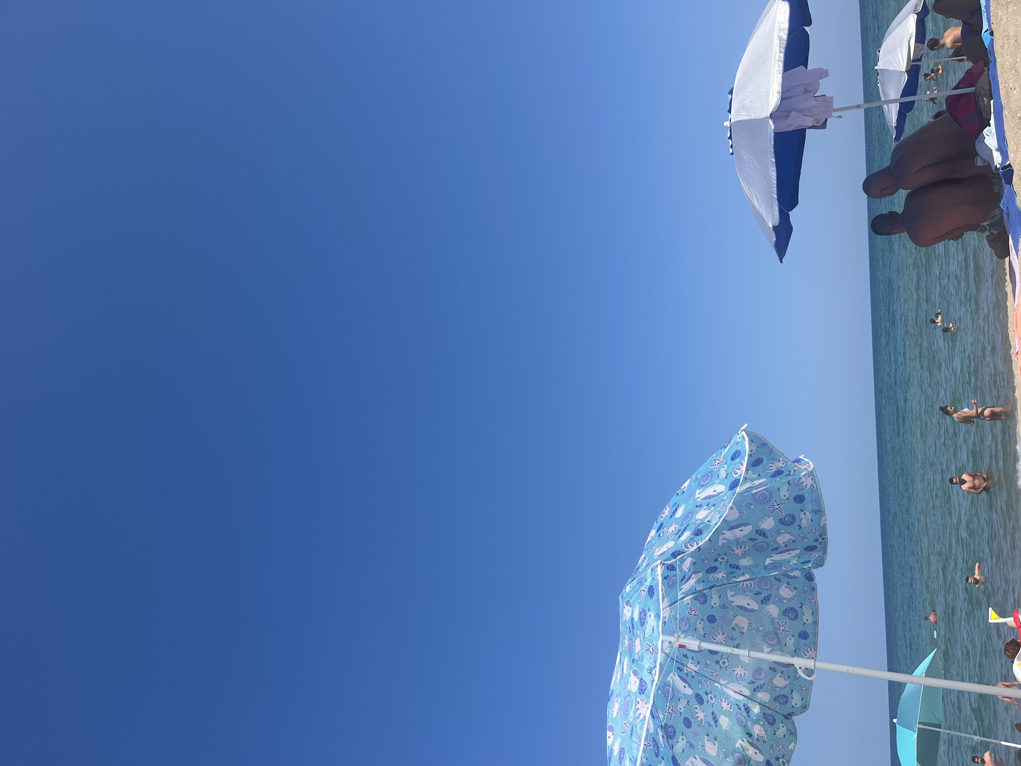 The image depicts a sunny day at the beach in Obzor, Bulgaria. The sky is a clear, vibrant blue, with a vast expanse above the calm, blue sea. In the foreground, two beach umbrellas provide shade. The umbrella on the left is light blue with a whimsical pattern of marine life, including octopuses, starfish, and shells. The umbrella on the right is white and blue, with white towels draped over its frame. Several people are visible in the shallow water and on the sandy shore. Some are swimming or wading, while others are sitting or standing under the umbrellas. In the middle ground, two men are sitting together under the right umbrella, facing away from the camera towards the sea. The overall scene conveys a relaxed atmosphere of a summer day at the beach.