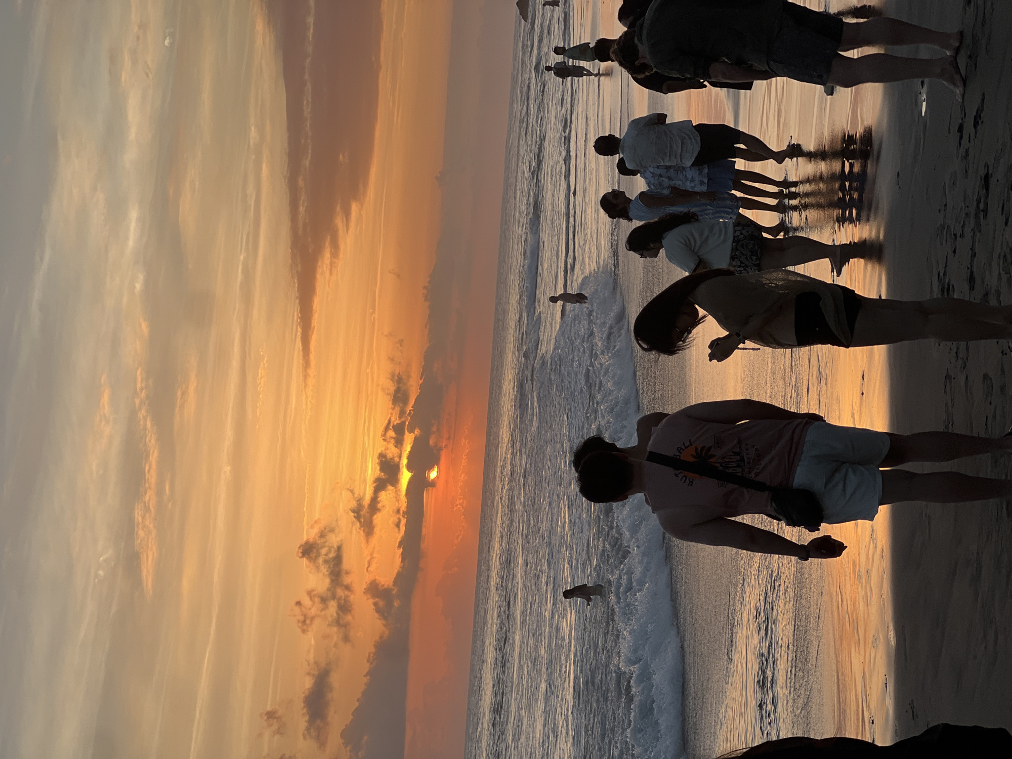 The image captures a sunset scene at a beach in North Kuta, Indonesia, with a group of people visible. The sky is ablaze with orange and yellow hues, indicating the sun is low on the horizon, partially obscured by clouds. The ocean has moderate waves, with some individuals wading in the water and others standing on the wet sand. In the foreground, several people are silhouetted against the bright sky and water. The sand is dark and reflects the sunset colors.