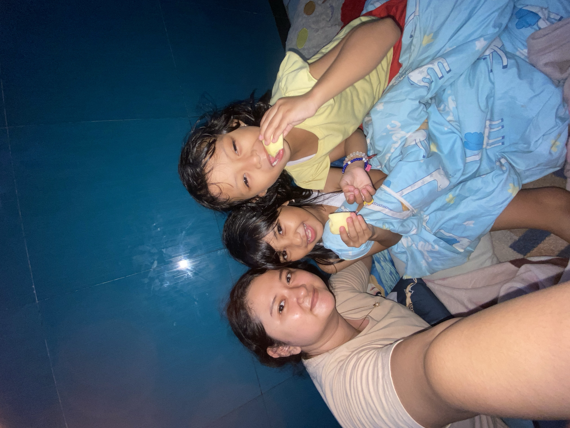 A selfie shows a woman with two girls, presumably her daughters, on a blue tiled floor. The woman is in the foreground, wearing a light brown t-shirt, and smiling. The girls are on top of her, nestled in a blue comforter with a cartoon pattern. The girl on top is wearing a yellow t-shirt, and the one below her is wearing a blue shirt. Both girls are holding pieces of food, perhaps fruit. The location is possibly inside a home in Gelam, Indonesia, indicated by the blue tiled floor, common in Indonesian homes. They appear to be in a playful, familial interaction.