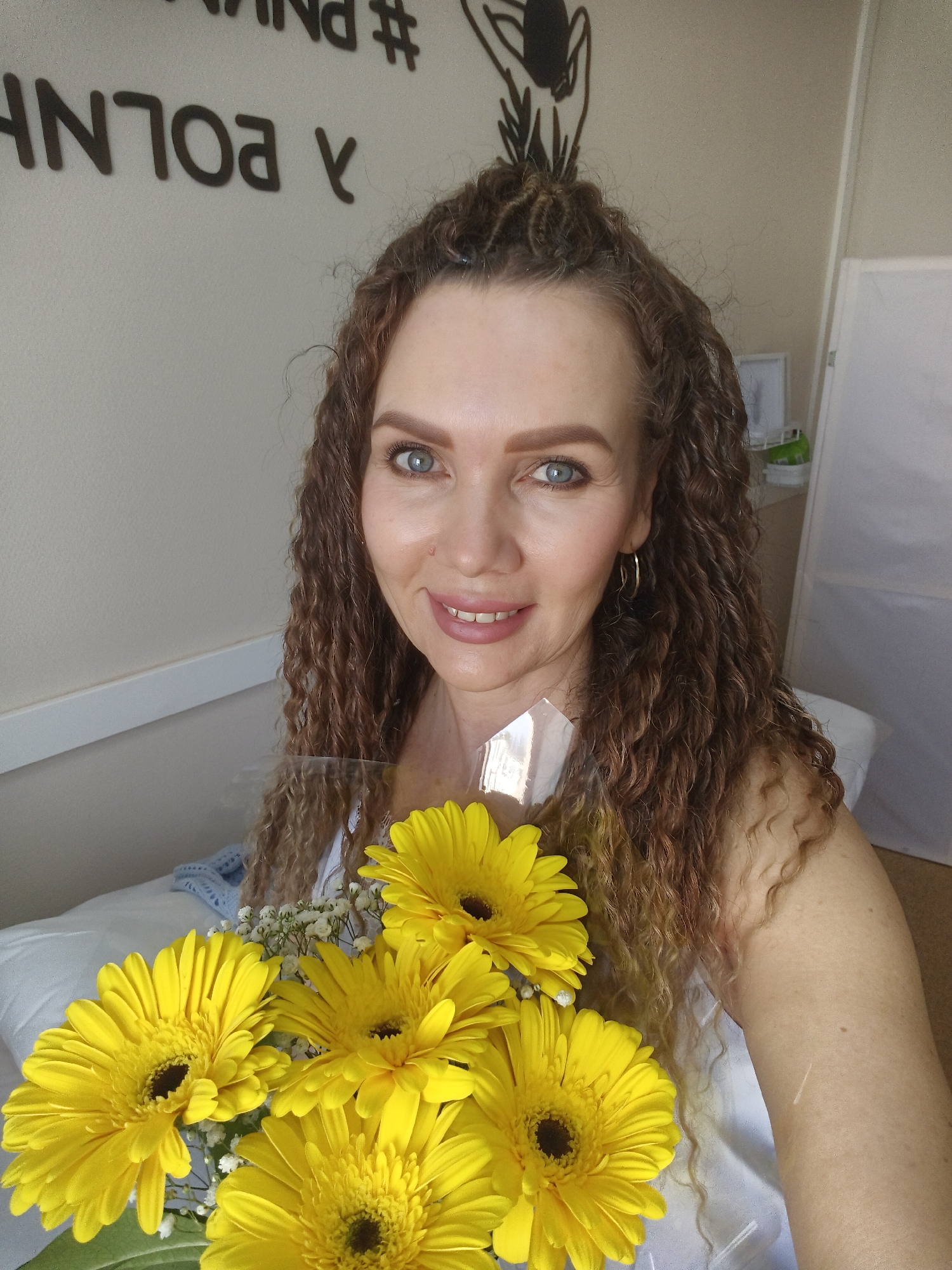 A medium shot depicts a woman holding a bouquet of yellow gerbera daisies and baby's breath. She has curly brown hair with some light brown highlights, blue eyes, and is wearing gold hoop earrings. A translucent plastic wrapping surrounds the bouquet. A white garment is visible under the flowers. The woman is positioned in what appears to be a room with a bed or padded surface covered with a white sheet or blanket. Behind her, on the wall, there is lettering. To the right of the woman, there is a white screen. The location is listed as Unknown, Unknown.