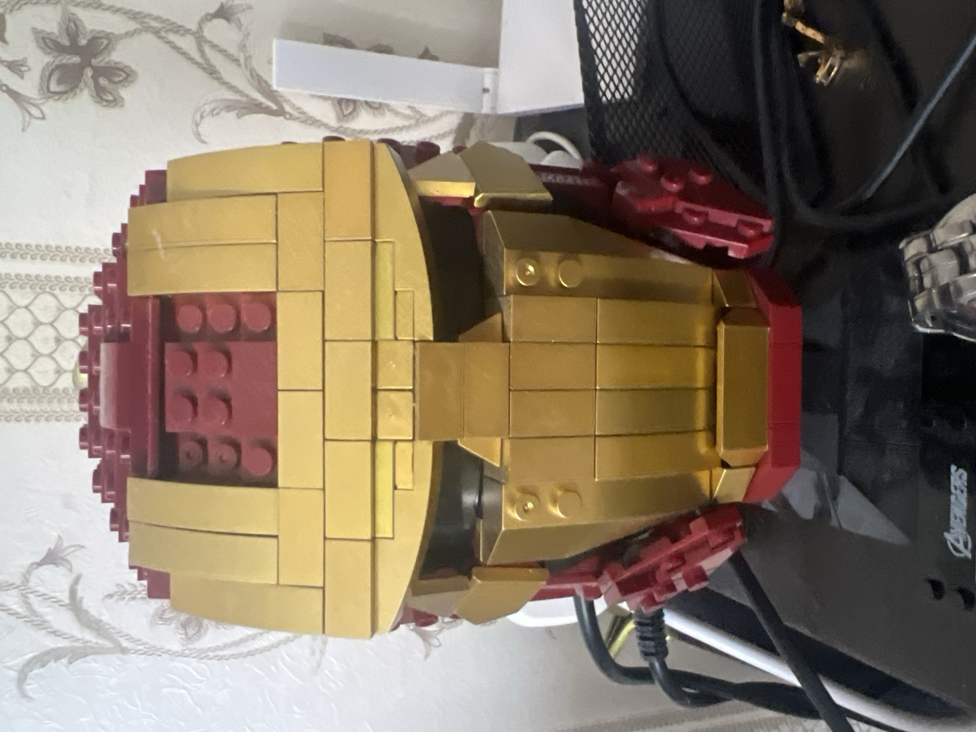 The image shows a close-up, head-on view of a LEGO Iron Man helmet, constructed with red and gold bricks. The helmet is placed on a black surface, which appears to be a desk or shelf. Behind the helmet, a wall with a patterned wallpaper is visible. To the left and right of the helmet, various cables and electronic devices are present, suggesting a home office or entertainment setup. The lighting is moderate, casting soft shadows. The scene is indoors, and no specific location within the city of Minsk, Belarus, can be identified from the visual information provided.