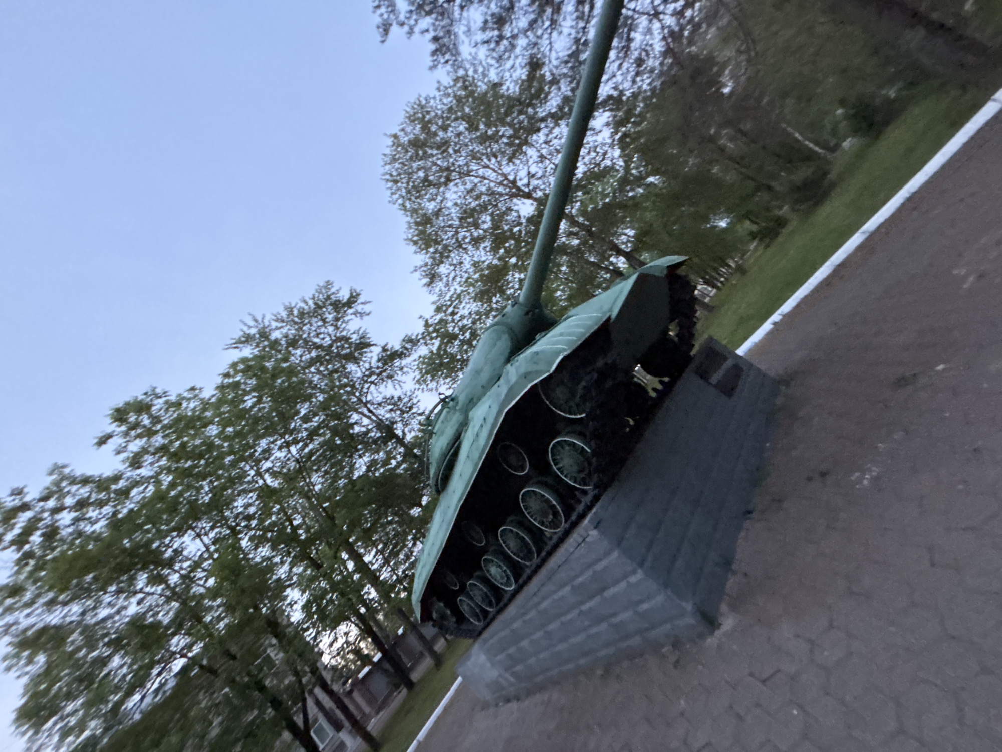 A low-angle, tilted photograph captures a green military tank mounted on a dark, rectangular pedestal in an outdoor setting. The tank, likely a monument, features visible tracks and road wheels, with its main gun barrel pointing upwards and to the left.
The scene is set under a light blue sky, suggesting either dawn or dusk due to the soft, muted lighting. Lush green trees with dense foliage surround the tank and form a backdrop, indicating spring or summer. Through the trees, a multi-story building with windows is faintly visible in the distance.
A paved area with an interlocking brick pattern is seen near the base of the pedestal. Further away, a darker path or road is bordered by a distinct white line or curb. No people or specific activities are depicted, and there is no visible text on the monument or surrounding elements. The overall impression is that of a quiet, commemorative space.