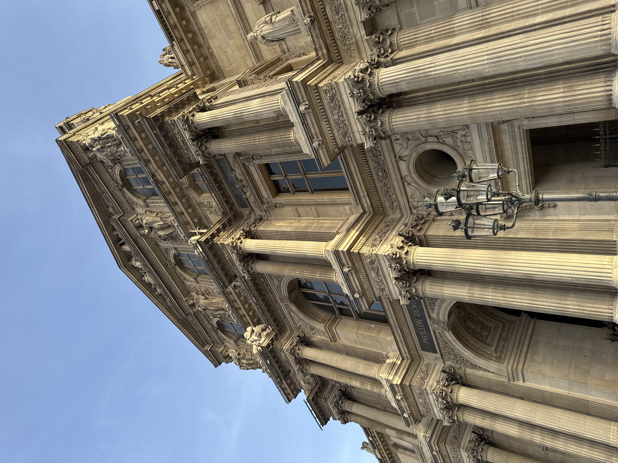The image shows an upward, tilted view of the Pavilion Denon at the Louvre Museum in Paris, France. The architecture is classical, featuring columns, windows, and ornate stone carvings. A vintage-style lamppost is affixed to the building. The sky is visible in the background, appearing blue.
