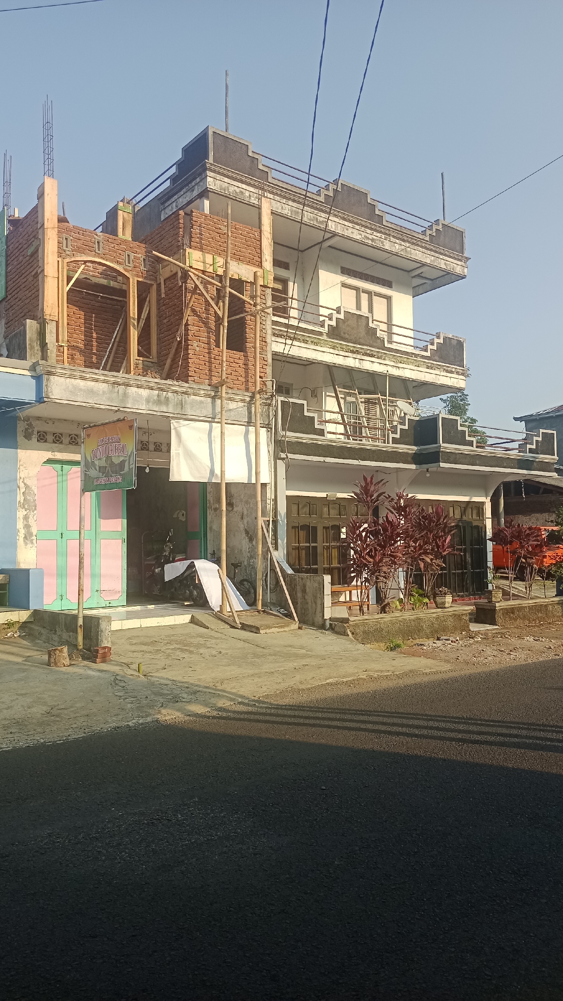 In the city of Karyamandala, Indonesia, a multi-story building is partially under construction and partially completed. The building features a white facade with dark decorative trim along the rooflines. On the left side, a section of the building is being built with exposed brick walls and wooden scaffolding. A banner hangs on the lower level of this section. The front of the building has storefront-like entrances with large glass windows. In front of one of these entrances are several red-leafed plants in planters. A paved road runs in front of the building, and shadows from the building and overhead wires are cast across it. The sky is clear and blue.