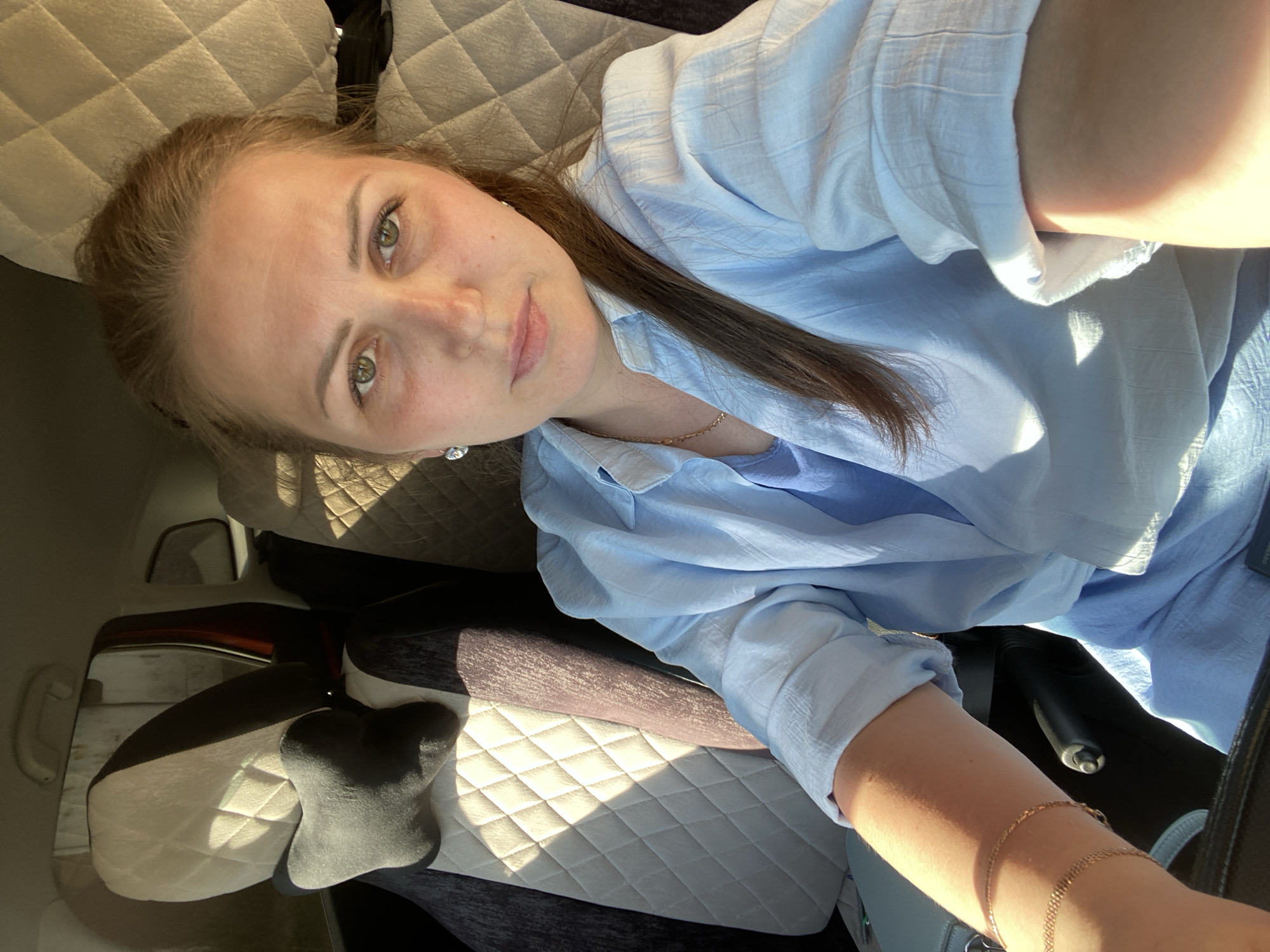 A woman with fair skin, brown hair, and green eyes is the main subject of the image. She is wearing a light blue button-down shirt and a delicate gold necklace and bracelet. She is in a vehicle with beige quilted seats. The image is taken from a high angle, looking down at the woman, likely from the front passenger seat. The location is unknown and cannot be determined from the image; the city of Markovo, Russia, is not identifiable within the visual data.