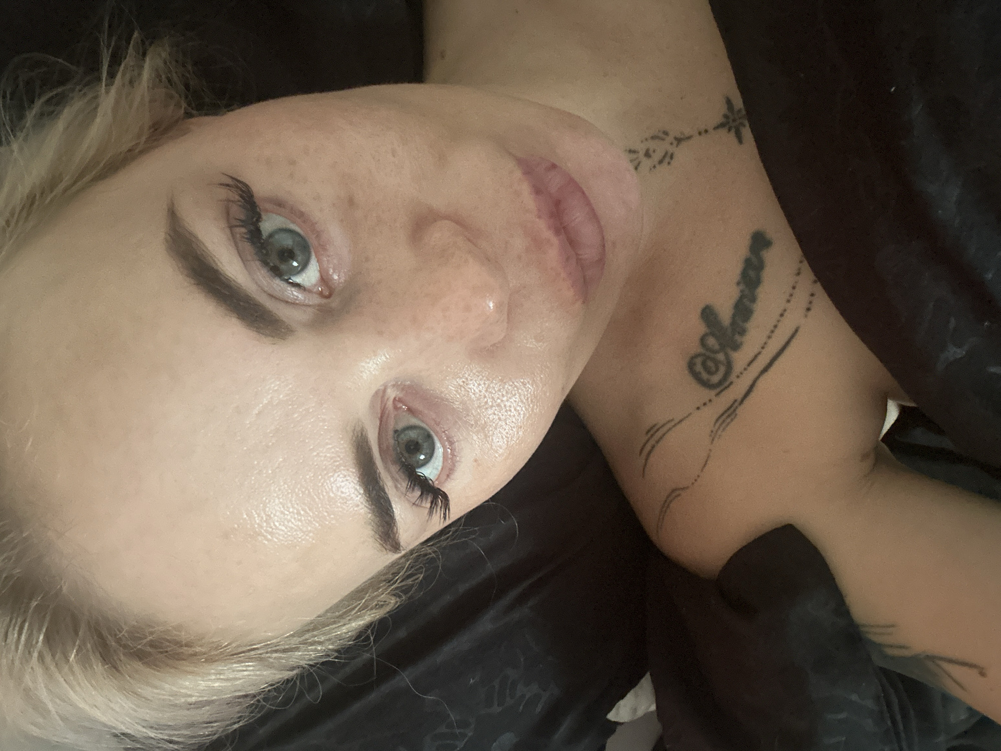 A close-up, top-down shot shows a woman with fair skin, freckles, and blonde hair. She is wearing false eyelashes and her makeup includes eyeliner and lip gloss. She has a tattoo of the word "Amara" on her left shoulder and chest, and another tattoo of a flower with a star on her upper chest. The woman is lying down, and the background is dark and appears to be fabric, possibly bedding. The image is in focus and well-lit.