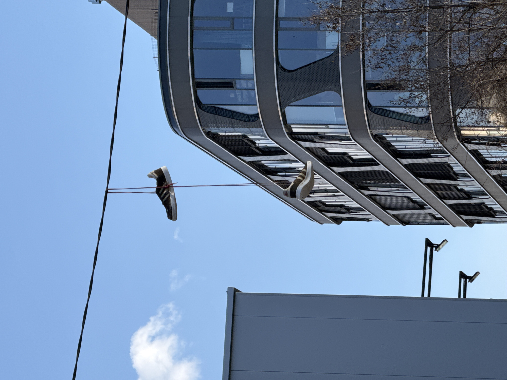 A pair of sneakers hang from a power line against a blue sky with scattered clouds, appearing to be tossed over the line with their laces tied together. The backdrop features a modern architectural building with a distinctive curved design and reflective glass windows, and a glimpse of another building beneath it. The location is Sofia, Bulgaria.