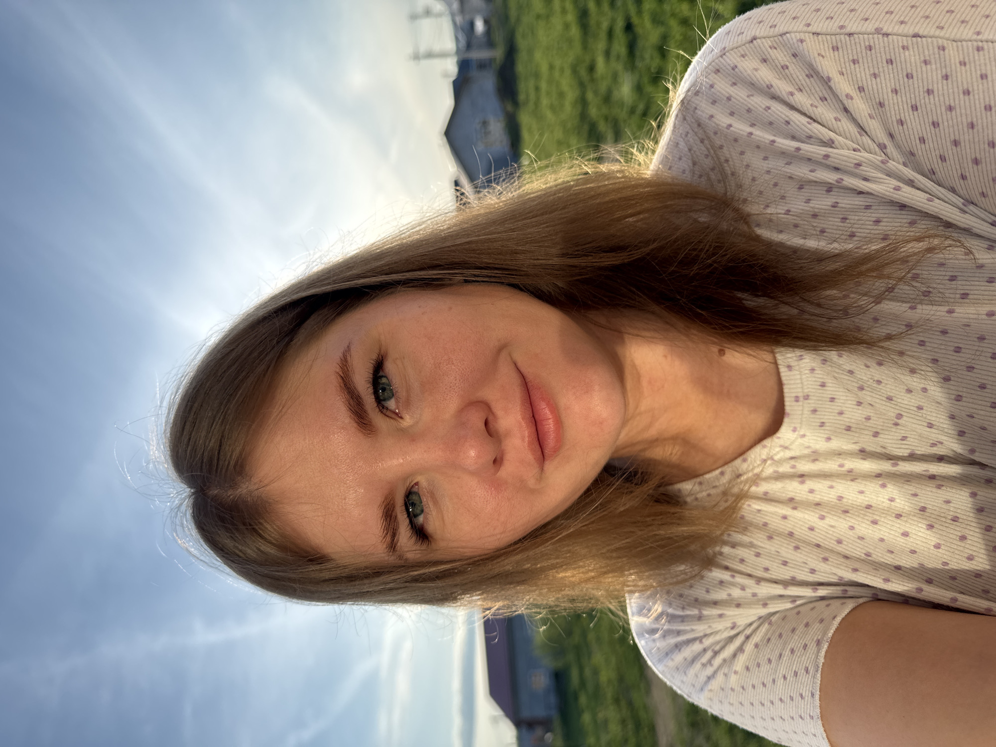 A young woman with fair skin and light brown hair is centered in the frame, her head slightly tilted to the right. She has green eyes and a neutral expression. She wears a white shirt with small lavender polka dots. The background consists of a blue sky with sunlight breaking through the clouds on the left side, and green grass along with a blurred building. The photo appears to be taken outside. Given the prompt's location, this scene is in Ферма, Russia.