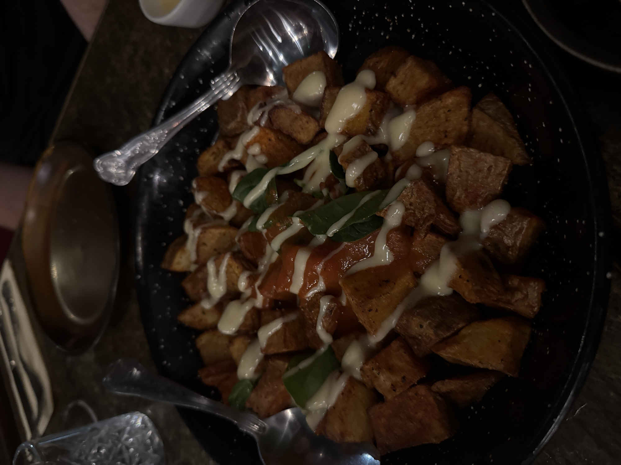 The image shows a plate of Patatas Bravas. The Patatas Bravas consist of fried potato cubes topped with a white garlic aioli and a red tomato sauce. Some fresh basil leaves are visible. The Patatas Bravas are served in a speckled black bowl or plate with two serving spoons. In the lower left corner is a drinking glass, and there's a white ramekin at the top of the image. Given the style of the dish, this photo was possibly taken in a restaurant or pub in Newcastle upon Tyne, United Kingdom.