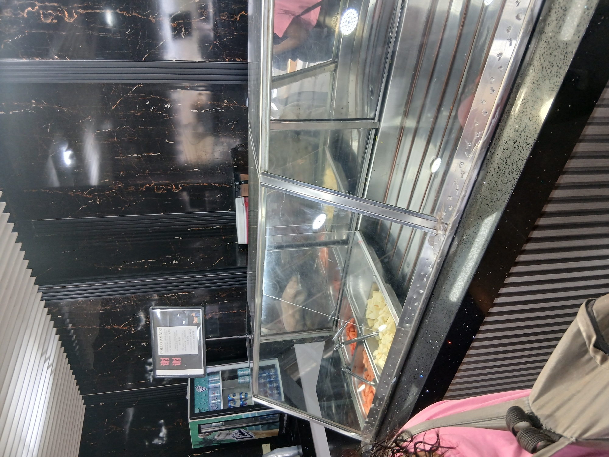 The image depicts the interior of a business establishment, likely a food vendor or restaurant, in Juan Griego, Venezuela. The scene is photographed from a high angle, showcasing a refrigerated display case filled with various prepared foods, including what appears to be chopped fruit and other items. The display case has a stainless-steel frame and glass panels.
The background features a wall with a dark, marble-like pattern, and another wall with alternating light and dark horizontal stripes. A menu board or sign is visible on the wall with the marble pattern. A cooler is partially visible beneath the food display case. A person wearing a pink shirt is partially in the image.