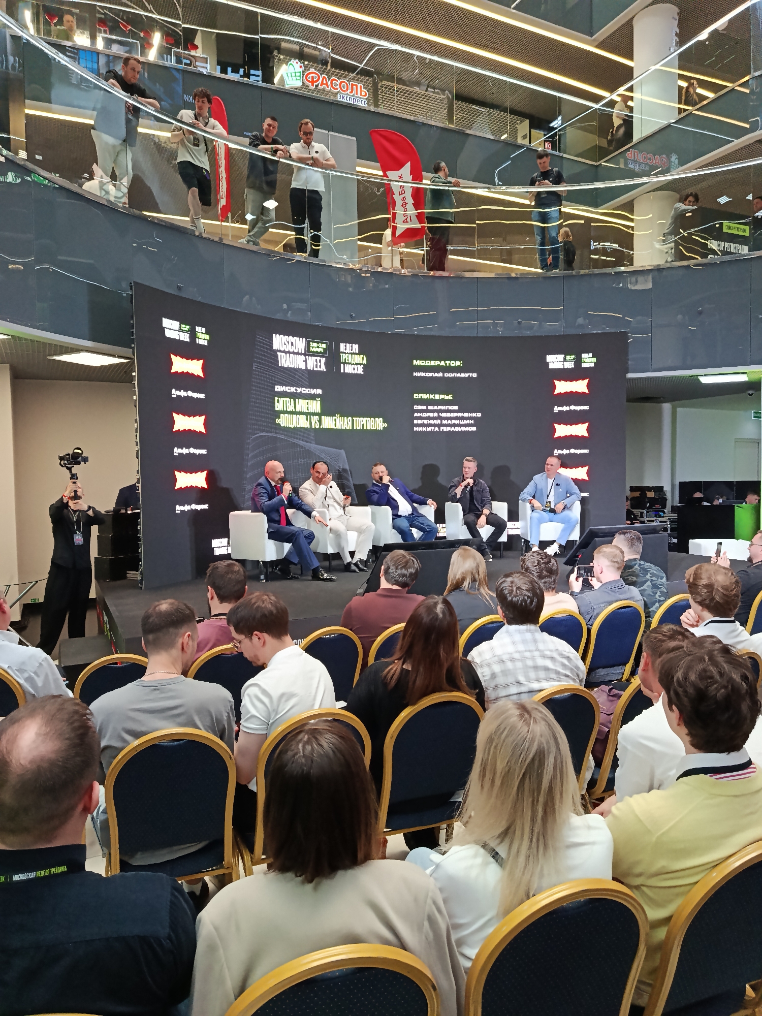 Here's a detailed description of the image:
**Scene:** An indoor event, likely a financial conference or seminar, taking place in what appears to be a multi-level shopping mall or large commercial building in Moscow, Russia. The primary focus is on a panel discussion.
**Setting:** A stage with a large screen displaying branding for "Moscow Trading Week" and the event's topic: "Battle of Opinions: Options VS Linear Trading" in Russian. The backdrop also features logos of "Alfa Forex" and "Alfa Bank". The environment is a modern shopping mall, featuring glass partitions, and metal infrastructure elements. Various shops are visible through the glass.
**People:**
* **Panelists:** Five men are seated on chairs on the stage, participating in a panel discussion. They are dressed in business casual or smart casual attire.
* **Moderator:** The man on the left, standing behind a camera, is likely filming the discussion.
* **Audience:** Attendees are seated in rows of chairs facing the stage. They are a mix of men and women, dressed in a variety of clothing styles. Some members of the audience are looking at their smartphones.
* **People on Balcony:** A group of people are standing on an upper level balcony or walkway, observing the event below.
**Objects:**
* Chairs arranged for the audience.
* The stage with the backdrop.
* Microphones for the panelists.
* A camera for recording the event.
* Branding materials and banners.
* Glass partitions on an upper level, suggesting a modern building design.
**Actions:**
* The panelists are engaged in a discussion.
* The audience is listening to the discussion.
* The camera operator is recording the event.
**Interactions:**
* The panelists are interacting with each other.
* The moderator is likely guiding the discussion.
* The audience is passively observing the event.
**Environment:**
* The event is taking place indoors, in a modern commercial building.
* The building has multiple levels and glass partitions.
* The overall atmosphere is professional and business-oriented.
