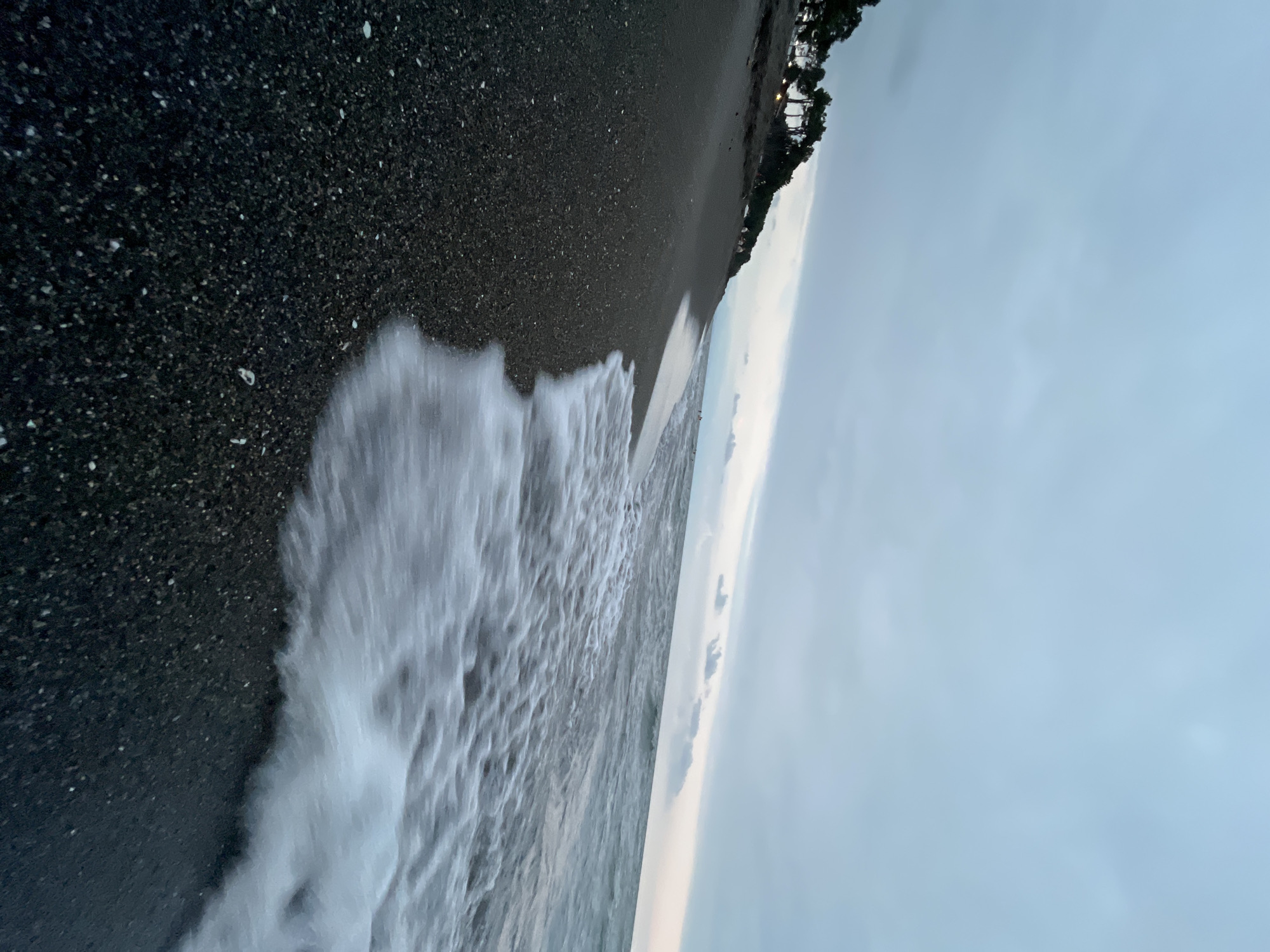 This is a low-angle shot of a black sand beach at Ureki, Georgia. The tide is coming in, with white foamy waves washing onto the dark, pebbly shore. In the background, a line of dark green trees runs along the top of a gentle incline, and the sky is overcast with muted grey and pale blue clouds. There are faint lights visible in the distance behind the trees, suggesting a populated area. A few distant figures can be seen near the treeline and further down the beach. The overall mood is tranquil and slightly moody due to the overcast sky and the dark sand.