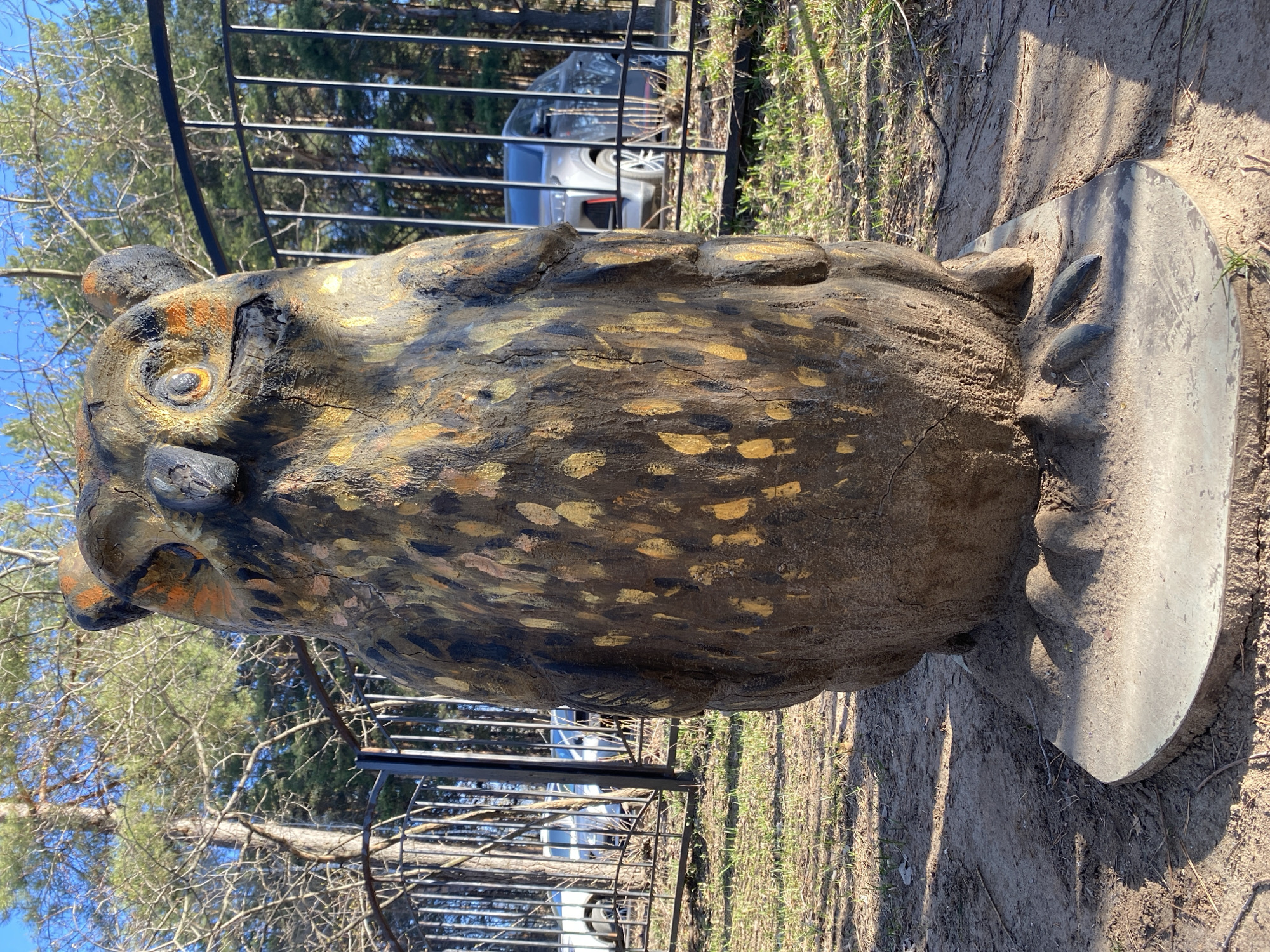 The image shows a wooden sculpture of an owl, standing on a circular concrete base. The sculpture is predominantly brown with golden-yellow painted spots. It has prominent eyes and features suggesting an owl's beak. The sculpture is situated outdoors, with a metal fence partially visible in the background. Through the fence, glimpses of vehicles can be seen. The surrounding ground is covered in dirt, with sparse grass. The trees and blue sky are visible above the fence. The subject matter and style align with folk art themes commonly found in public spaces, such as parks or gardens. The location is Chaikovsky, Russia.