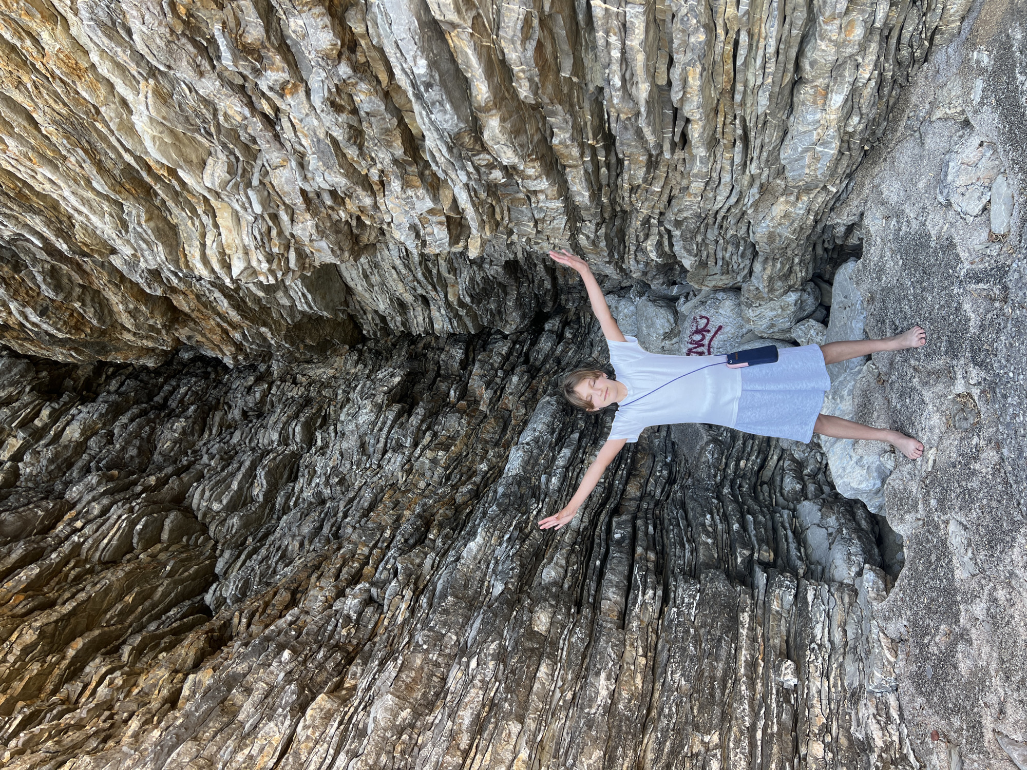 A person is lying on a rock formation in an outdoor setting. The person is fair-skinned with short hair, wearing a white shirt and light-colored dress. A thin strap hangs across their chest. Their arms are outstretched. The rock formation is layered. The environment appears to be a natural, rocky area. The location is Budva, Montenegro.