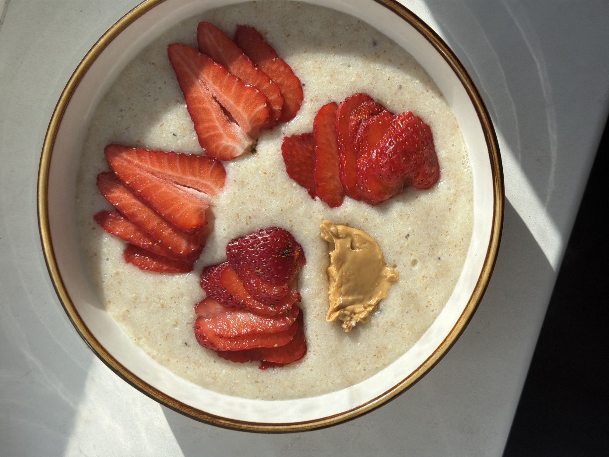 A top-down, close-up shot reveals a white bowl with a brown rim. Inside the bowl is oatmeal, topped with several piles of sliced strawberries and a dollop of peanut butter. The bowl sits on a white surface. The lighting suggests a bright, daytime setting. The location is not discernible from the image.