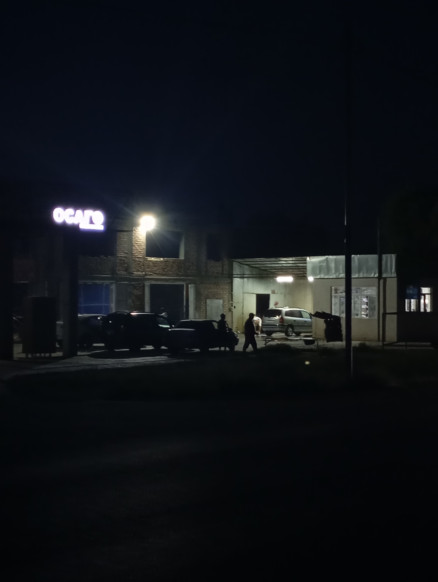 The image depicts a nighttime scene in an urban setting, likely a commercial area. In the background, a building with exposed brickwork and large window openings is visible. A prominent illuminated sign with the text "OGATO" in large, purple, stylized letters is mounted on the left side of the building. A bright overhead light shines from the upper right of the building. In front of the building, several vehicles are parked, including sedans and what appears to be an SUV. Two individuals are visible near the vehicles: one is standing in the foreground, facing away from the camera, and another is walking towards the right of the frame. The ground in front of the building is paved, and there is a dark, unlit area in the foreground, suggesting a street or parking lot. The overall atmosphere is one of a business or service establishment operating after dark. The location is identified as Kızıl-Bel, Kyrgyzstan.