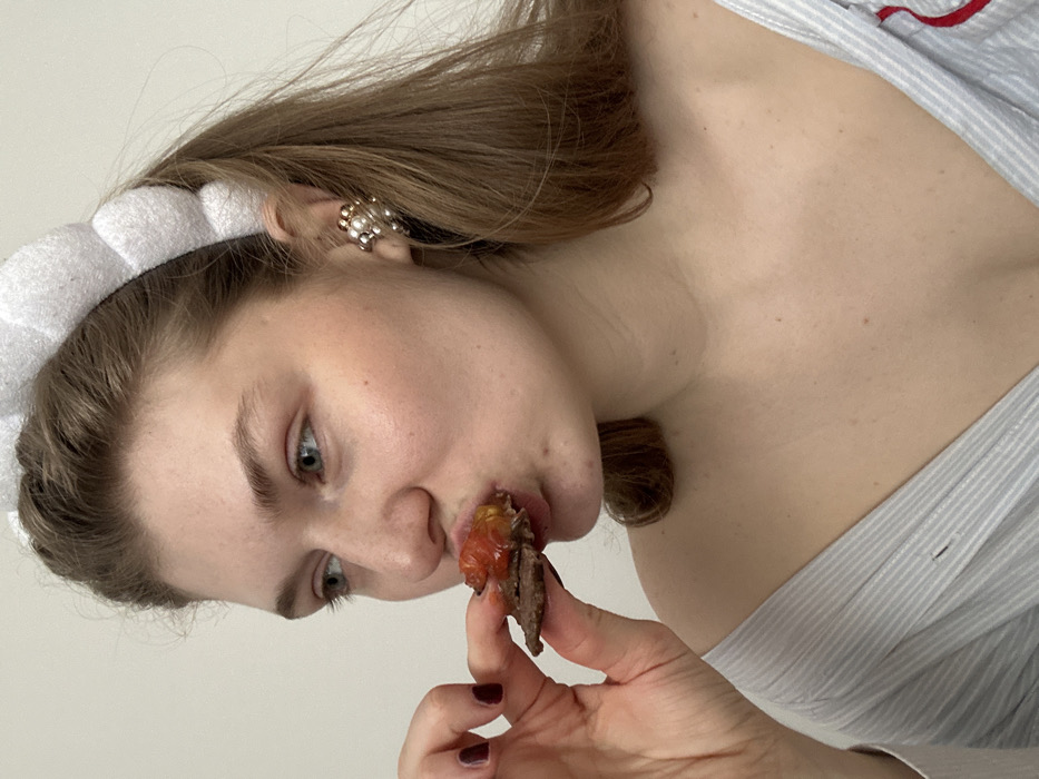 The scene, located indoors in Matiushino, Russia, depicts a young adult female from a close perspective. She has light brown hair and green eyes. She is wearing a white, segmented headband and a light-colored, off-the-shoulder garment with blue and white stripes; a red strap is visible on the garment's right side. A clustered earring featuring multiple pearls and gold-toned accents is present on her right ear. Her left hand, adorned with dark red nail polish, holds a small food item consisting of a dark, solid base topped with a reddish, semi-viscous substance. The subject is in the process of consuming this item, with her mouth partially open around it. The background consists of a plain, light-toned wall.