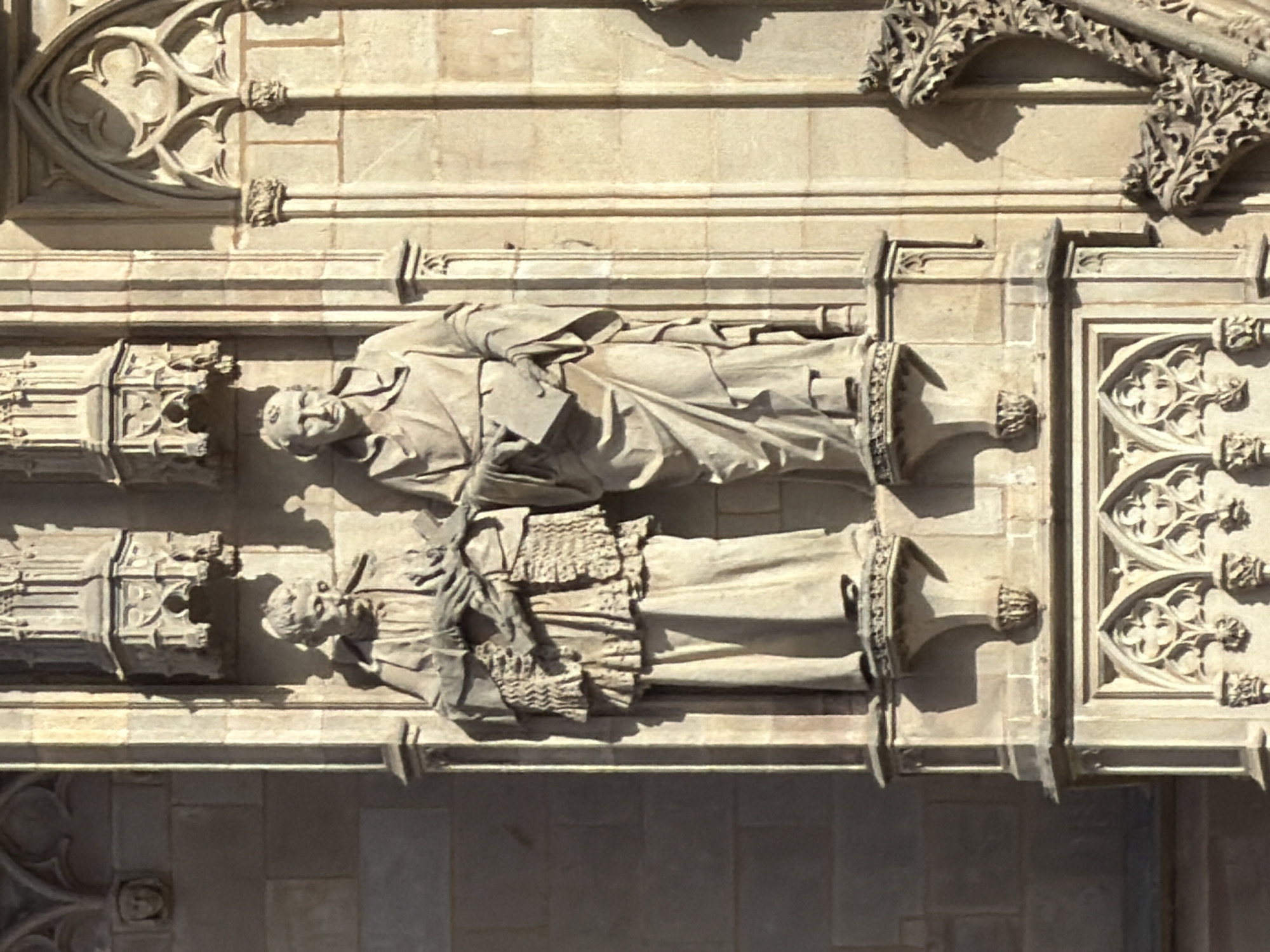 The image depicts a close-up view of architectural sculptures on the facade of a building in Barcelona, Spain. Two stone statues of robed figures are positioned side-by-side. The figure on the left is holding a book in their hands, which are crossed over their chest, and is wearing a layered, ornate garment. The figure on the right is also holding a book and wearing a draped robe. The statues are mounted on a stone facade with Gothic architectural elements, including decorative carvings and fluted columns. The lighting suggests it is daytime, with strong shadows cast by the architectural features and the statues.
