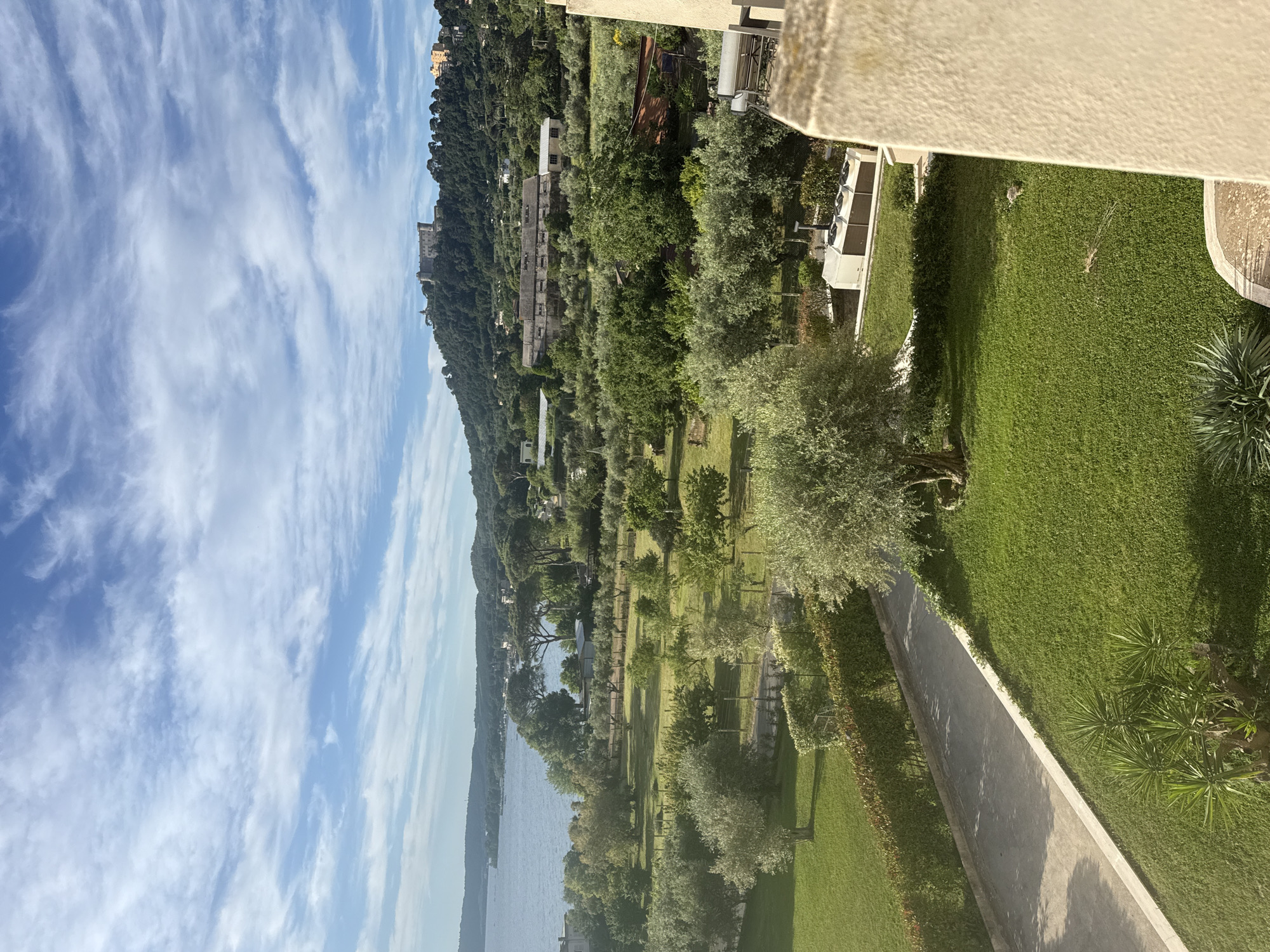 The image presents a landscape in Bracciano, Italy, captured from an elevated vantage point, likely a balcony or high window.

The scene comprises a body of water (likely Lake Bracciano) to the left, bordered by lush greenery with trees and lawn leading to the lake. A well-maintained lawn is to the right. The sky is partly cloudy with streaks of clouds that suggest windy conditions.

In the distance is a hill covered with buildings amidst the trees. One structure stands out as a historical building.

The foreground shows details of the building where the photo was taken, including a concrete balcony railing, adding depth to the composition. A paved path or walkway leads to the property.