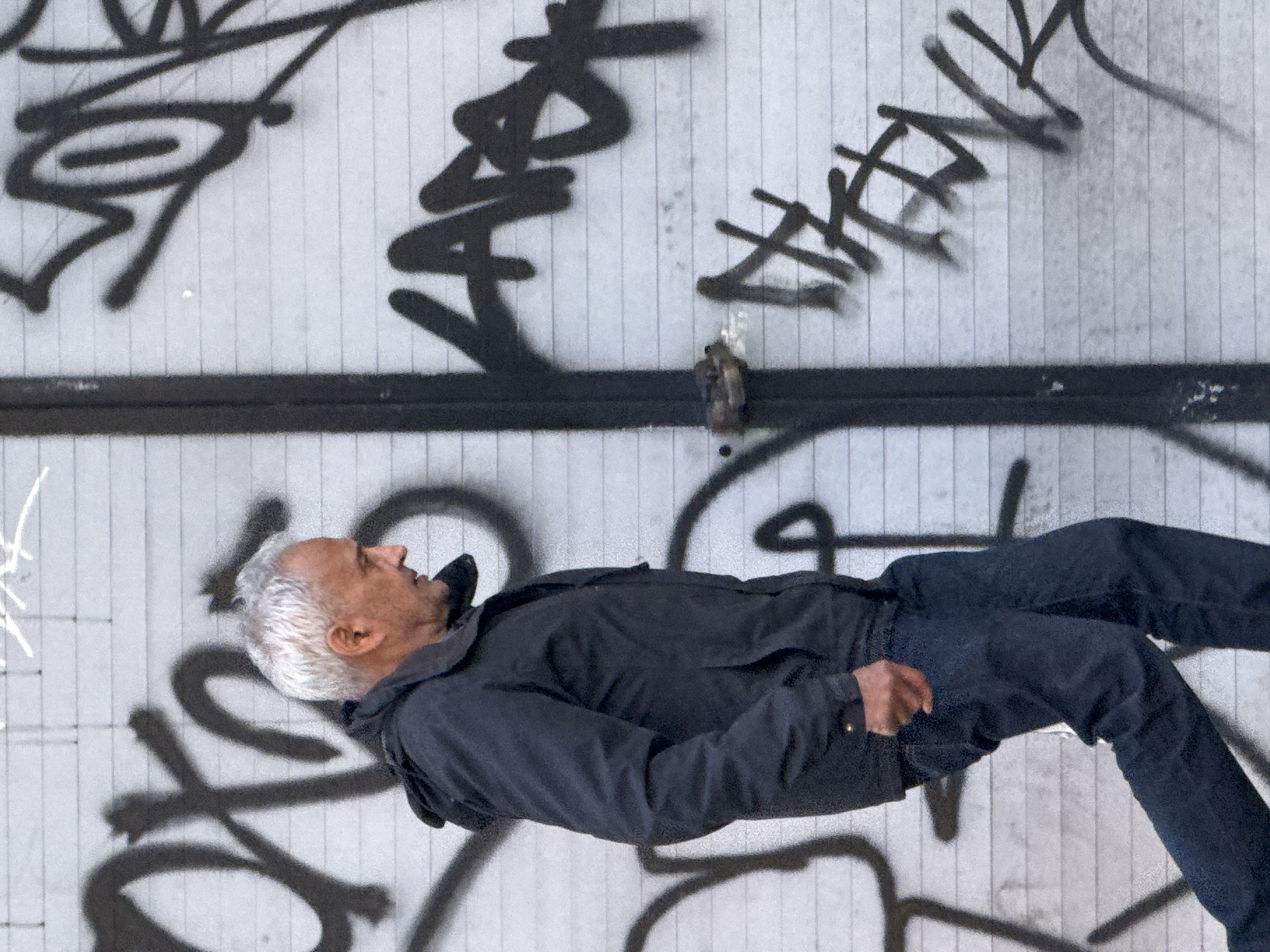 A man with short, white hair lies on his back against a gray wall covered in black graffiti. He wears a dark-colored jacket and jeans. The wall appears to be corrugated with vertical lines. A metal fixture is visible near the top center of the wall. The exact location in London, United Kingdom is not determinable from the image.