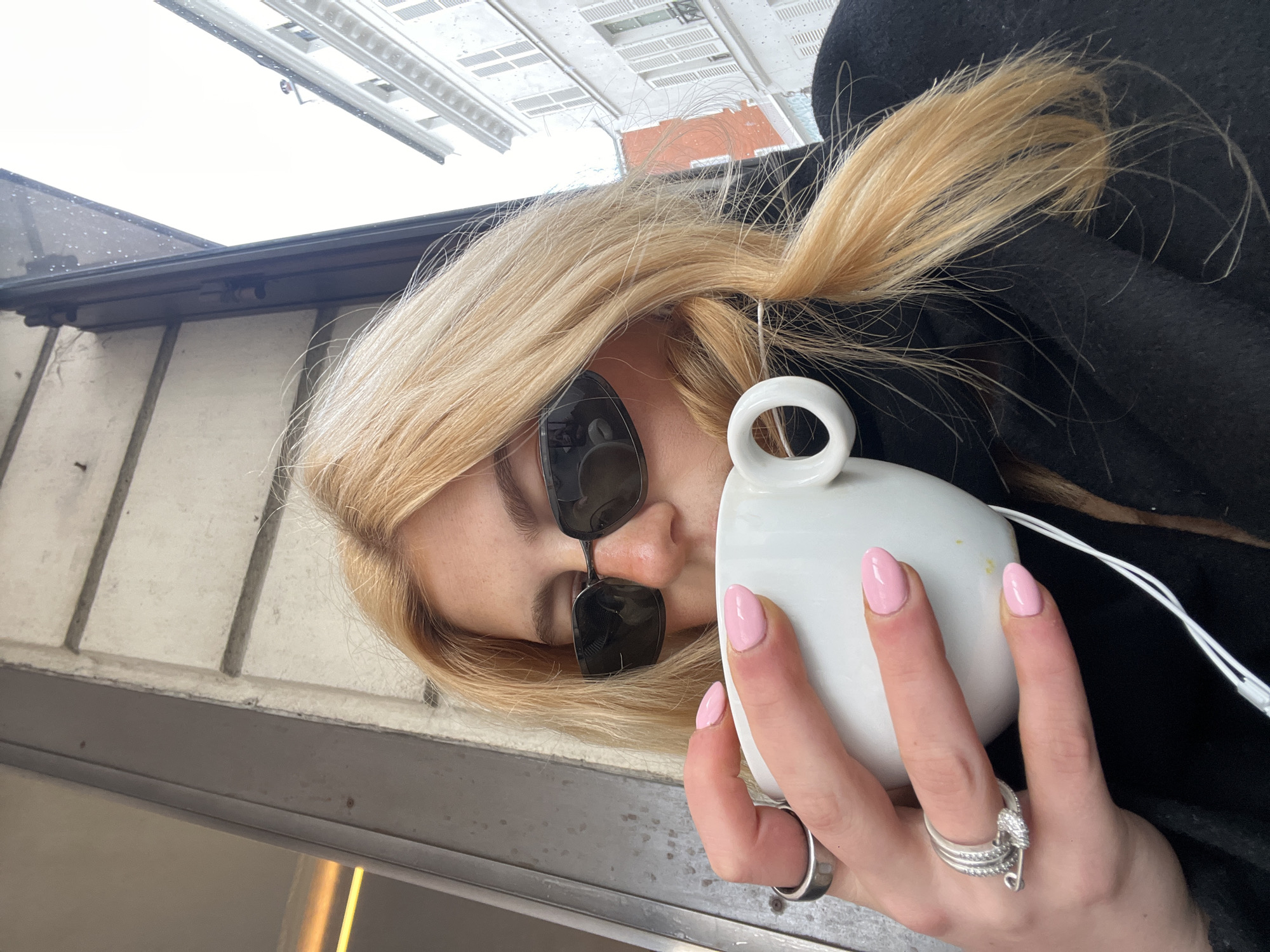 The image depicts a person with blonde hair, wearing dark rectangular sunglasses and a black outer garment, drinking from a white mug held with both hands. The individual's left hand is adorned with pink nail polish, a thick silver band on the index finger, and multiple silver rings on the ring finger, one resembling a safety pin design. A white wired earbud is visible near the person's right shoulder, with the wire extending downward. The background features typical Haussmannian architecture characteristic of Paris, France, including multi-story buildings with light-colored facades and white shutters. An overhead awning or structure exhibits small water droplets, indicating precipitation or recent rain under an overcast sky. The scene is located in the city of Paris, France.