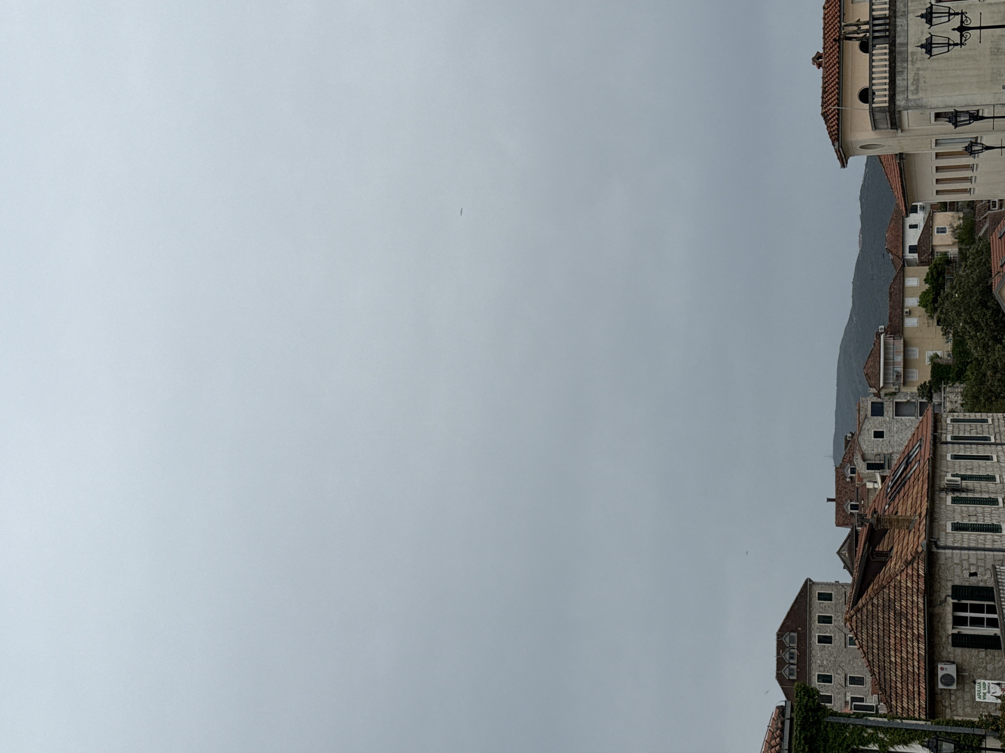 The image, rotated counter-clockwise 90 degrees, shows a cloudy sky above the buildings in the city of Herceg Novi, Montenegro. The buildings are of varying heights and architectural styles, with red tiled roofs and light-colored walls. Some buildings are newer than others. Behind the buildings is a hill or mountain. The cloudy sky takes up most of the image.