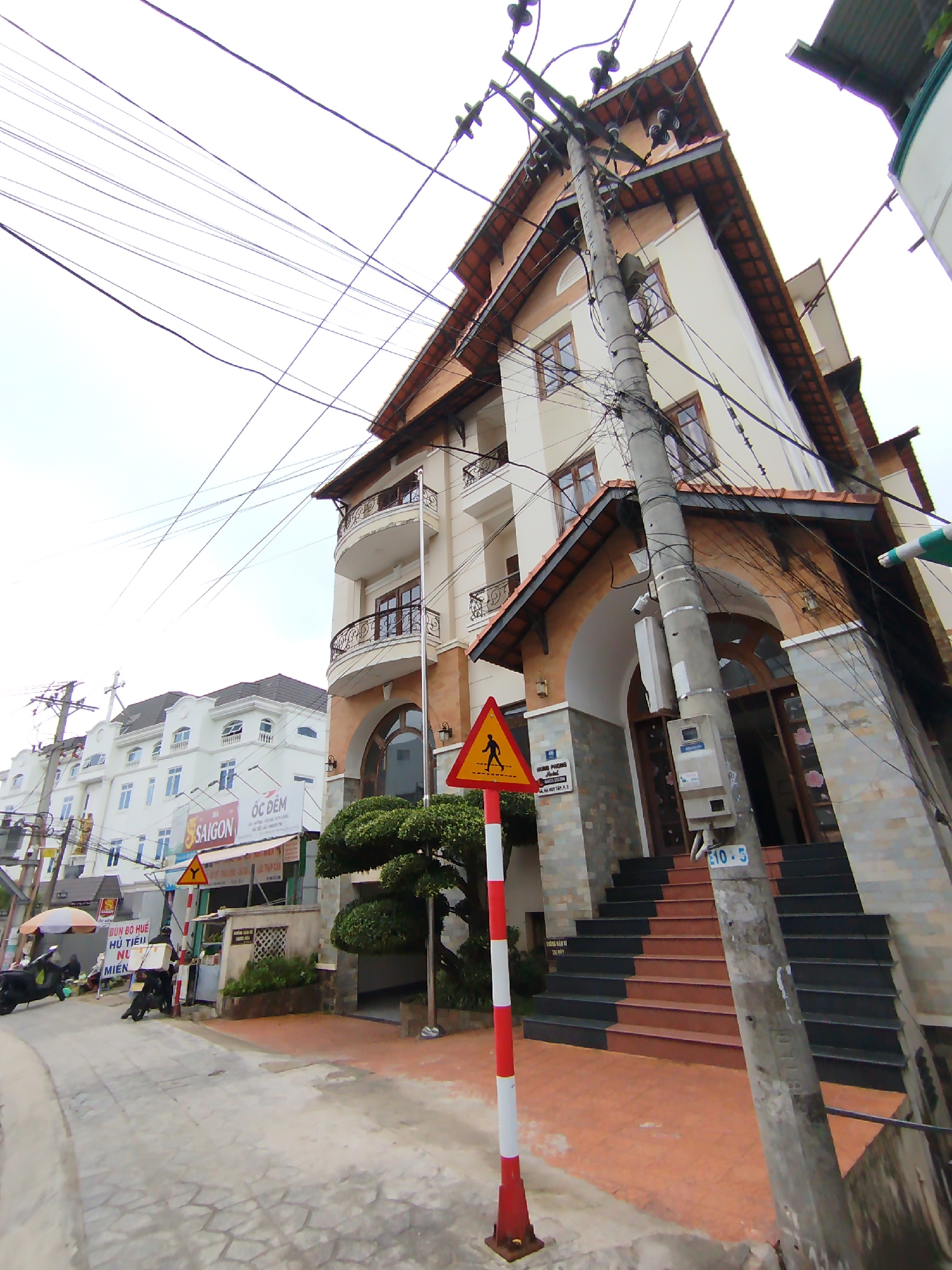 This image captures a vibrant street scene in Da Lat, Vietnam, under an overcast daytime sky. The perspective is looking up a slightly curving street towards a row of buildings.
In the immediate foreground and dominating the right side is a multi-story building, likely a hotel or residential complex, featuring a cream-colored facade, reddish-brown tiled roof, and intricate dark brown balcony railings. Its entrance is marked by a large archway and a set of dark and red-brown stairs leading up from the street level. A neatly trimmed green bush is planted near the entrance. A concrete utility pole heavily laden with a dense tangle of black electrical and communication wires stands prominently in front of this building. A red and white striped pole with a yellow triangular pedestrian warning sign (a black walking figure) is also visible in the foreground. A blue label "E10-5" is affixed to the utility pole.
Further down the street on the left, several other buildings are visible. Most notable is a large white building with multiple windows, a brown roof, and a visible cross on its rooftop, suggesting it might be a church or religious establishment. Signs are mounted on the facade of this building, including "SAIGON" in red, "ÓC ĐÊM" in black, and below that, text partially visible as "BÚN BÒ HUẾ HỦ TIẾU MIEK," indicating a food stall or restaurant.
On the street, near the food establishment, a person on a black scooter is seen, and another scooter is parked. There is also a vendor's stall with an umbrella.
Text visible on a sign on the main building's entrance reads "46 HOÀNG PHƯƠNG ANH ĐÀO" and "Ô BA HÚT TÂM 3." The street surface is paved with gray interlocking bricks.