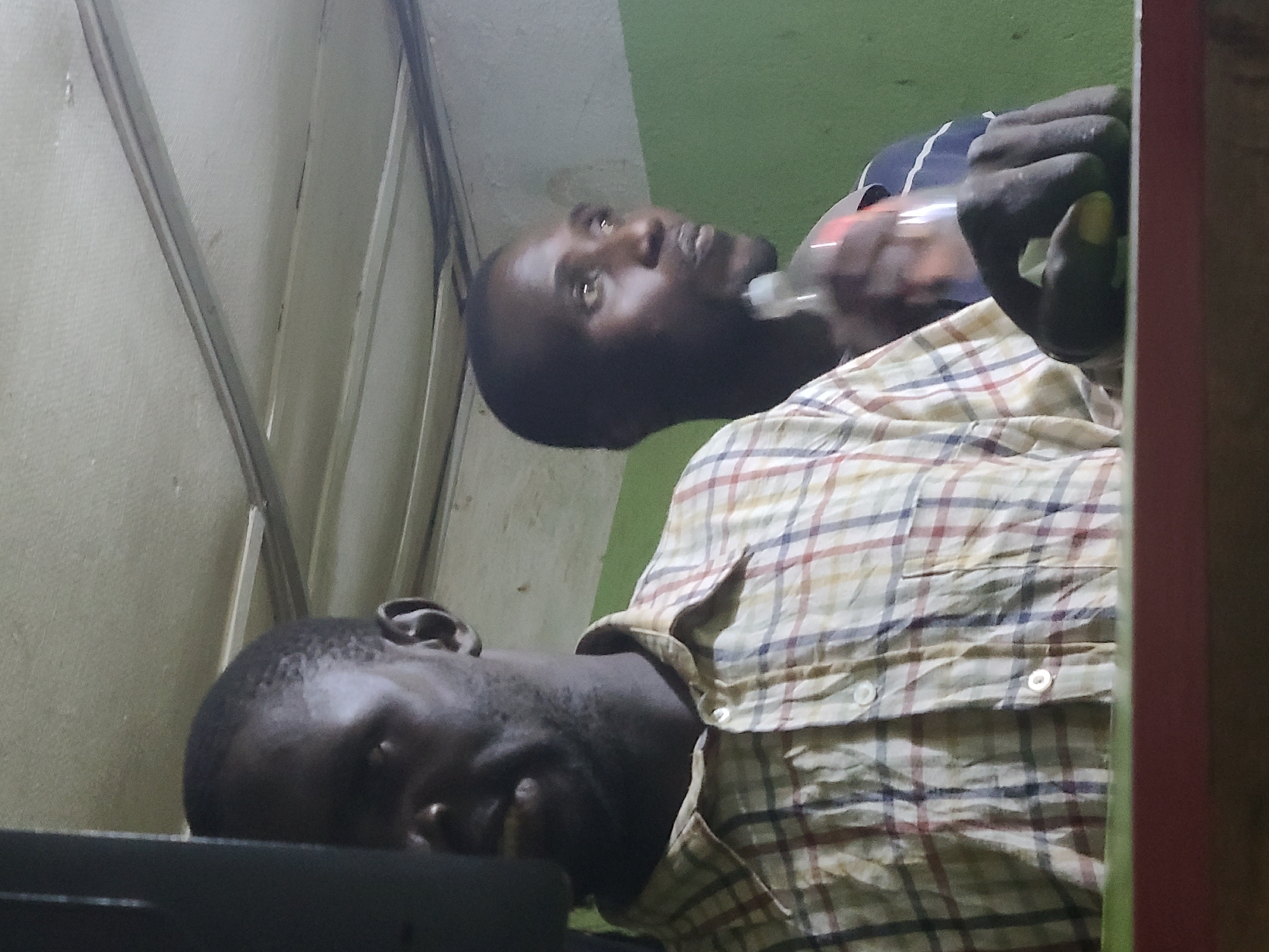 Two dark-skinned men are in an indoor space. One man is smiling, and part of his face and shoulder is visible. He wears a patterned collared shirt. The second man is further back, looking upward and holding a clear plastic bottle in his right hand. He is partially wearing a dark-colored t-shirt. The environment features walls of off-white and green, with metal frame pieces visible in the upper left. The exact location within Jos, Nigeria is not determinable from the visual data.