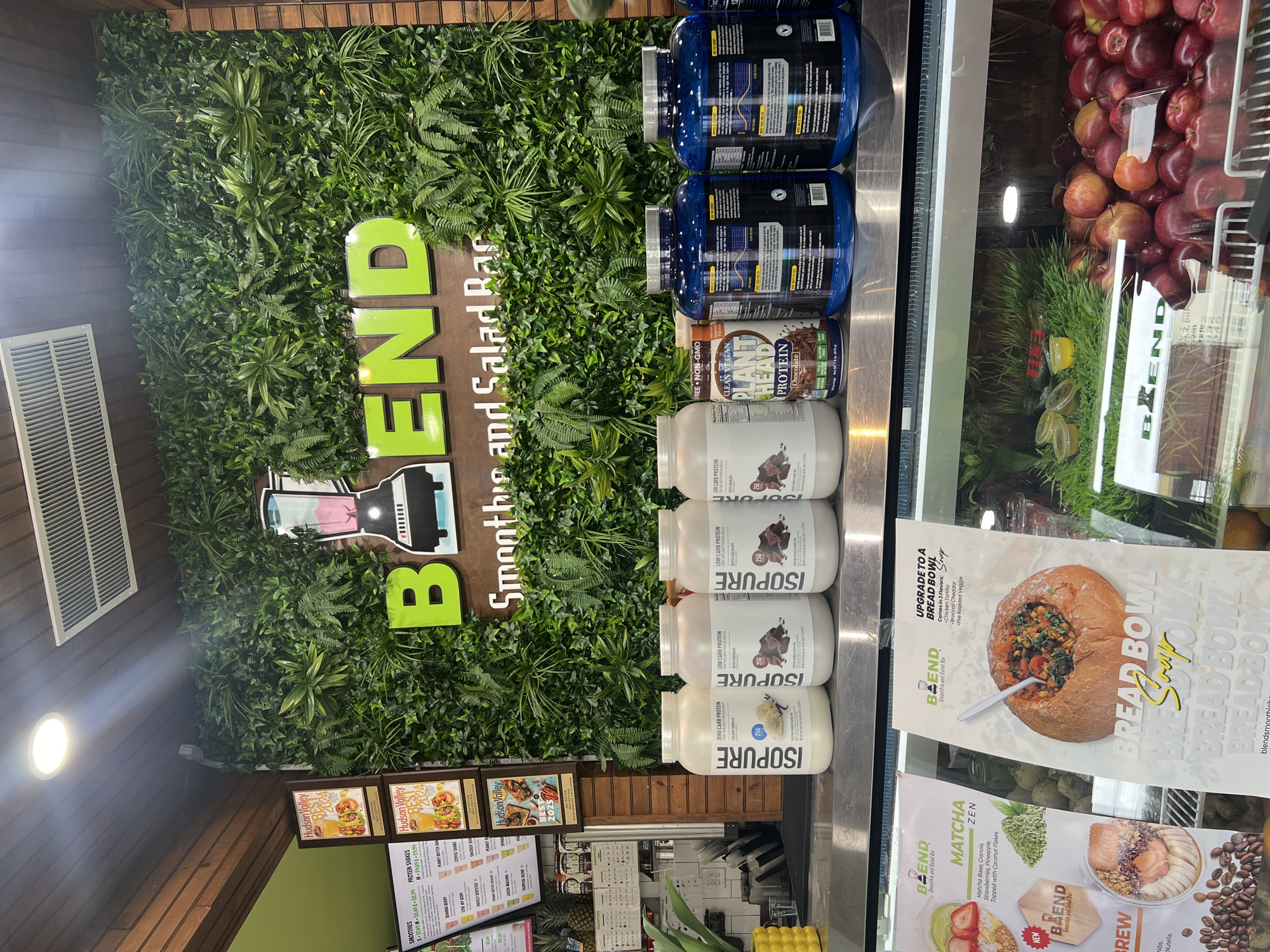 Here is a detailed description of the image:
**Scene:** The image depicts the interior of a "Blend" Smoothie and Salad Bar, located in an unknown city in the Town of New Windsor, United States of America. The establishment has a fresh and health-conscious vibe.
**Signage and Decor:**
- A large sign reading "BLEND, Smoothies and Salad Bar" is prominently displayed against a wall covered in artificial green foliage. The word "BLEND" is in green, and the rest of the text is in brown on a wooden-looking background.
- A logo featuring a silhouette of a blender is incorporated into the signage.
- There are additional promotional posters for menu items like Hudson Valley 2024.
**Menu and Products:**
- A menu board lists various smoothie and protein shake options, along with their prices.
- Shelves or a refrigerated display are stocked with protein powders, including brands like "ISOPURE" and "CLEAN VEGAN PROTEIN."
- There are fresh apples and other ingredients in a glass display case.
- Promotional material for "BREAD BOWL Soup" and "MATCHA ZEN" is displayed.
**Overall Impression:** The image conveys a sense of a modern and health-focused eatery offering a variety of smoothies, salads, and protein supplements.