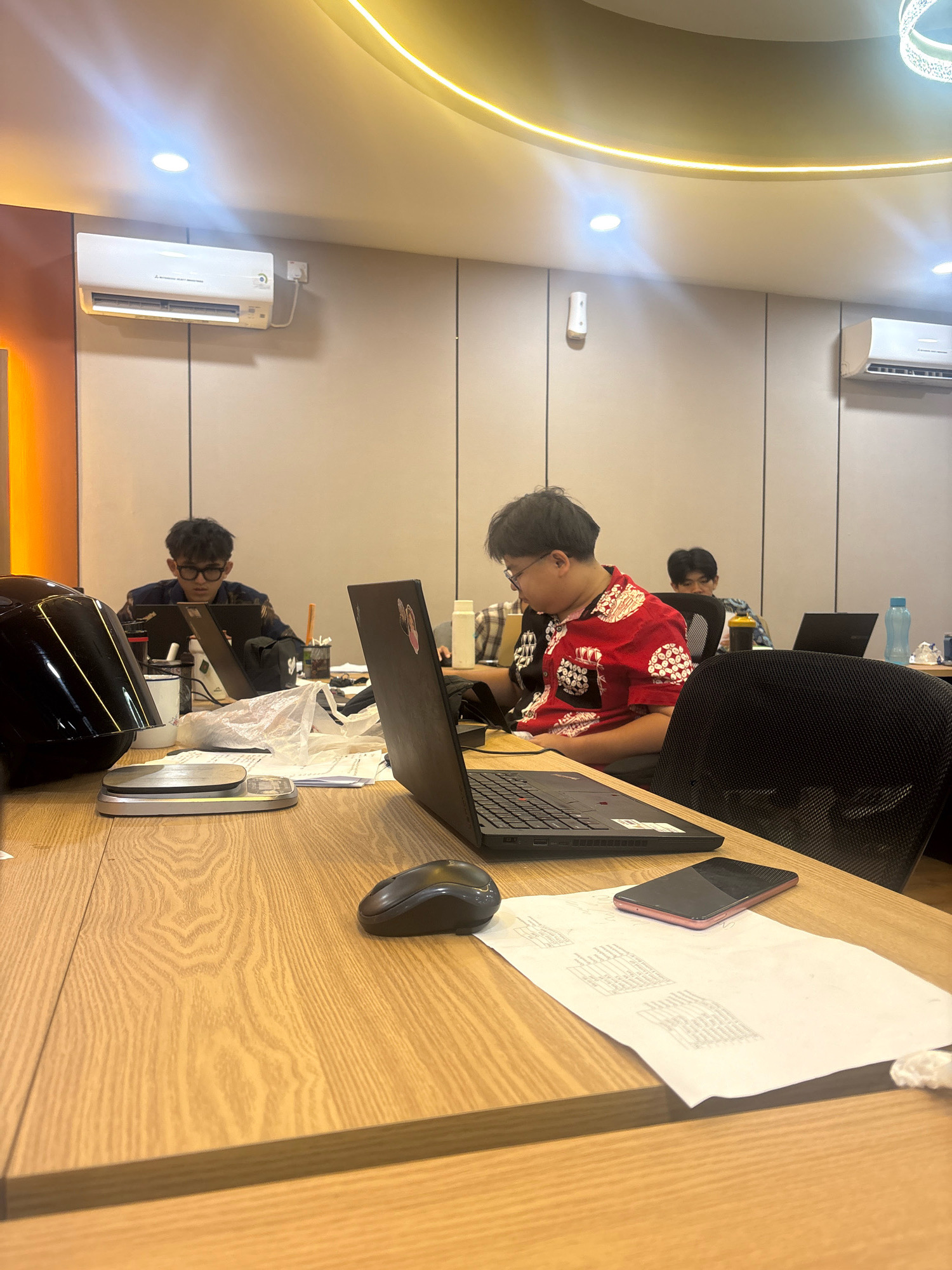 The image depicts an indoor scene, likely an office environment in Batam, Indonesia, with several individuals working at desks. The room features light-colored, paneled walls and a modern ceiling with integrated lighting and a decorative circular light fixture. Two air conditioning units are visible mounted on the walls.
In the foreground, a wooden desk is prominently featured. On the desk, a laptop is open, and next to it, a computer mouse, a pink smartphone, and a stack of papers are visible. Further back, a person wearing a red and white patterned shirt is seated at a desk, also engaged with a laptop. To the left, another individual wearing glasses and a dark jacket is working on a laptop. In the background, a third person is seated at a desk, and a blue water bottle is on a desk. The overall impression is one of a functional workspace.